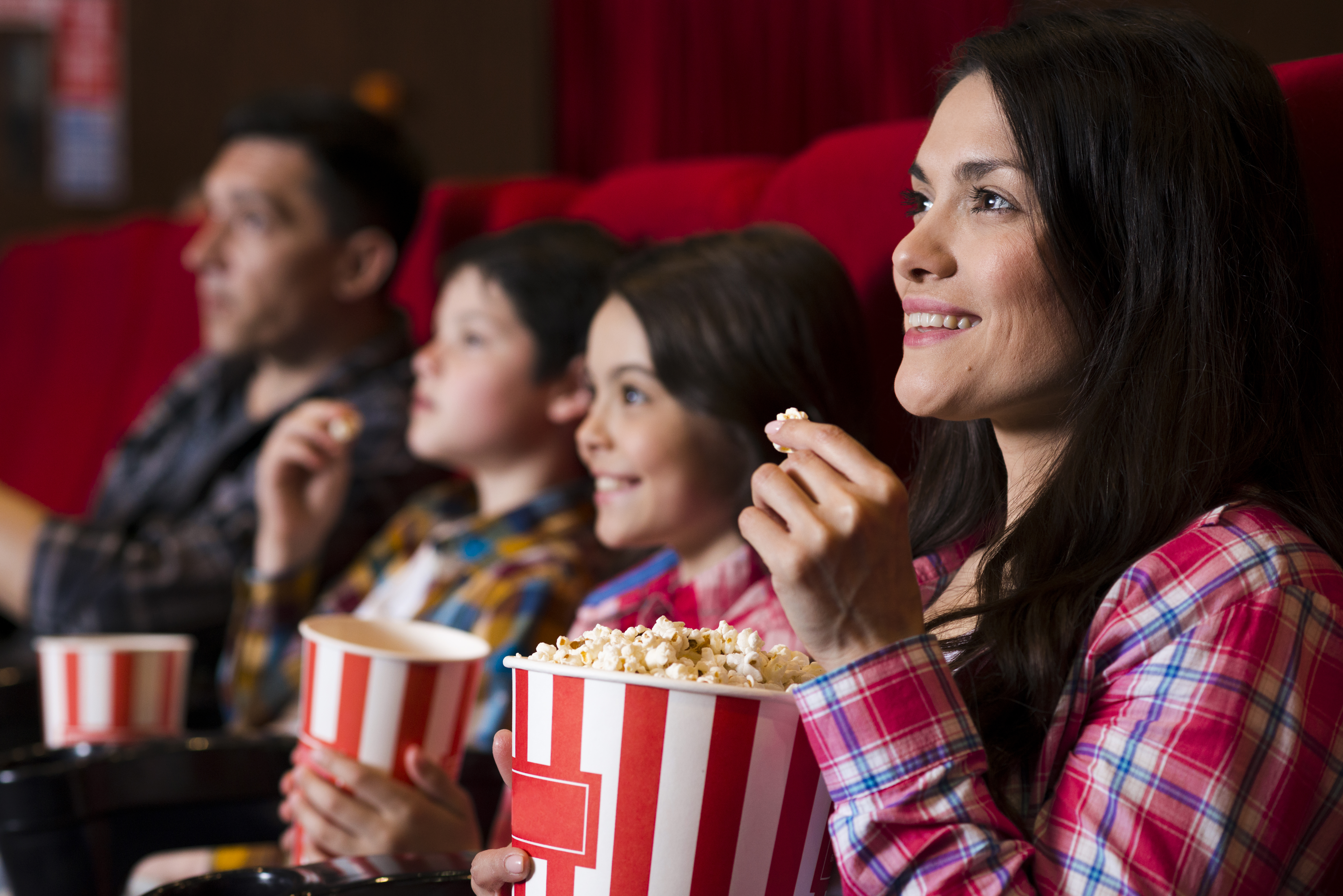 Fam&iacute;lia assistindo a um filme no cinema, com a m&atilde;e sorrindo e comendo pipoca de um balde listrado vermelho e branco. Duas crian&ccedil;as e um homem est&atilde;o sentados ao lado, tamb&eacute;m com pipoca, olhando para a tela