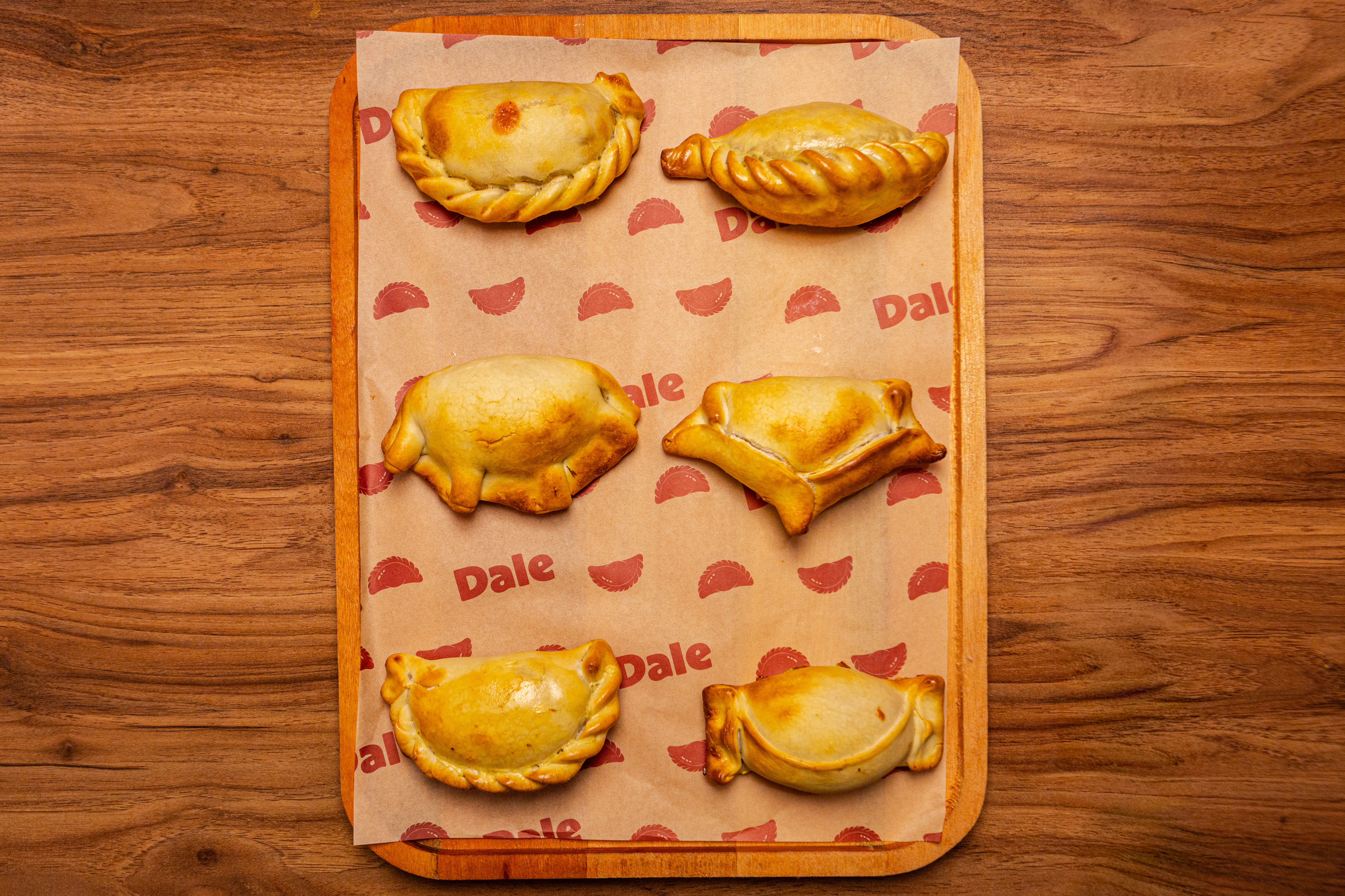 Seis empanadas assadas, douradas, com bordas repuxadas e formatos variados, sobre papel manteiga estampado com o nome Dale e pequenas empanadas vermelhas, dispostas em uma bandeja de madeira clara sobre uma mesa de madeira escura