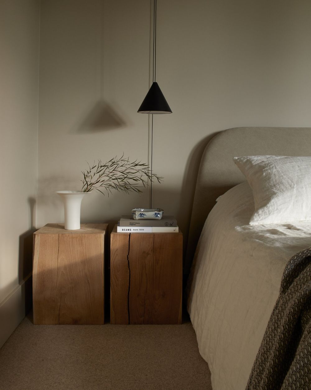 Quarto com cama de casal e cabeceira estofada bege. Duas mesas de cabeceira de madeira r&uacute;stica, uma com vaso branco e galhos secos, outra com livros e um cinzeiro decorado. Um pendente preto em formato de cone ilumina o ambiente. O ch&atilde;o &eacute; coberto por carpete bege e a parede &eacute; cinza-esverdeada