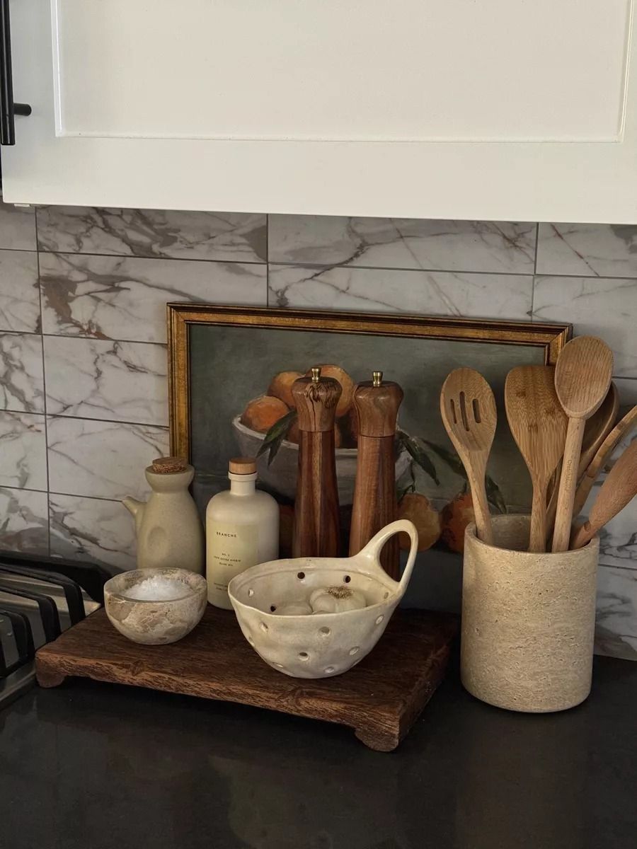 Bancada de cozinha com azulejos de m&aacute;rmore cinza e branco, arm&aacute;rio superior branco. Sobre uma t&aacute;bua de madeira r&uacute;stica, h&aacute; um saleiro, um recipiente de cer&acirc;mica com alho e duas garrafas de azeite. Ao fundo, um quadro com pintura de frutas c&iacute;tricas. &Agrave; direita, um pote de cer&acirc;mica com utens&iacute;lios de madeira