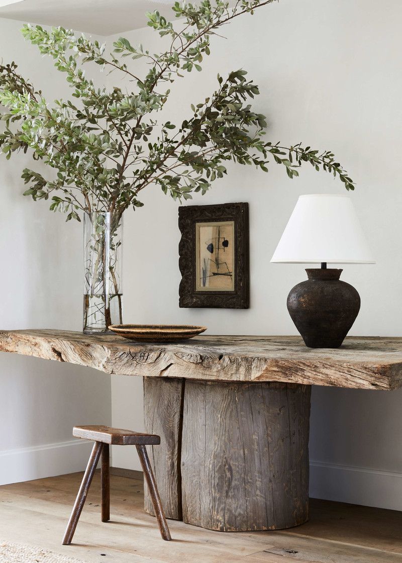 Mesa r&uacute;stica de madeira maci&ccedil;a com tampo irregular e base cil&iacute;ndrica, decorada com vaso de vidro com galhos verdes, um quadro abstrato emoldurado e um abajur com base escura e c&uacute;pula branca. Um banquinho de madeira de tr&ecirc;s pernas est&aacute; &agrave; esquerda, no ch&atilde;o de madeira clara