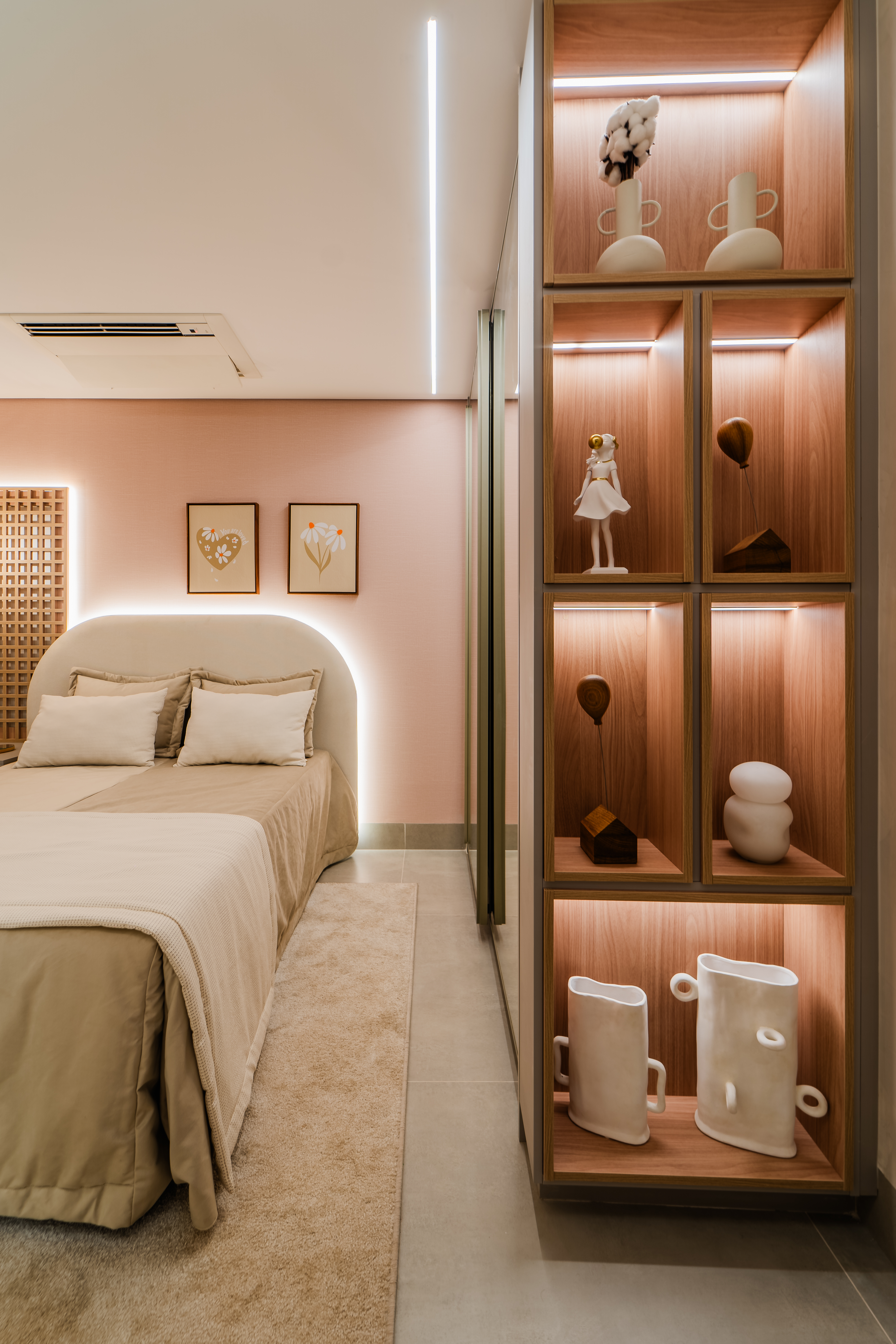 Quarto moderno com paredes rosa claro, cama de casal bege com cabeceira estofada e ilumina&ccedil;&atilde;o indireta. &Agrave; direita, uma estante de madeira clara com nichos iluminados, exibindo objetos decorativos brancos e marrons. No ch&atilde;o, um tapete bege ao lado da cama.