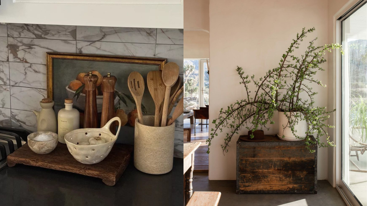 Cozinha e sala com decoração rústica e tons neutros. À esquerda, bancada com utensílios de madeira, saleiro, azeite e alho em potes de cerâmica, sobre uma tábua de madeira. Ao fundo, azulejos de mármore e um quadro com frutas. À direita, um vaso branco com planta de galhos finos e folhas verdes, sobre um baú de madeira escura, ao lado de uma janela grande que revela um ambiente externo claro