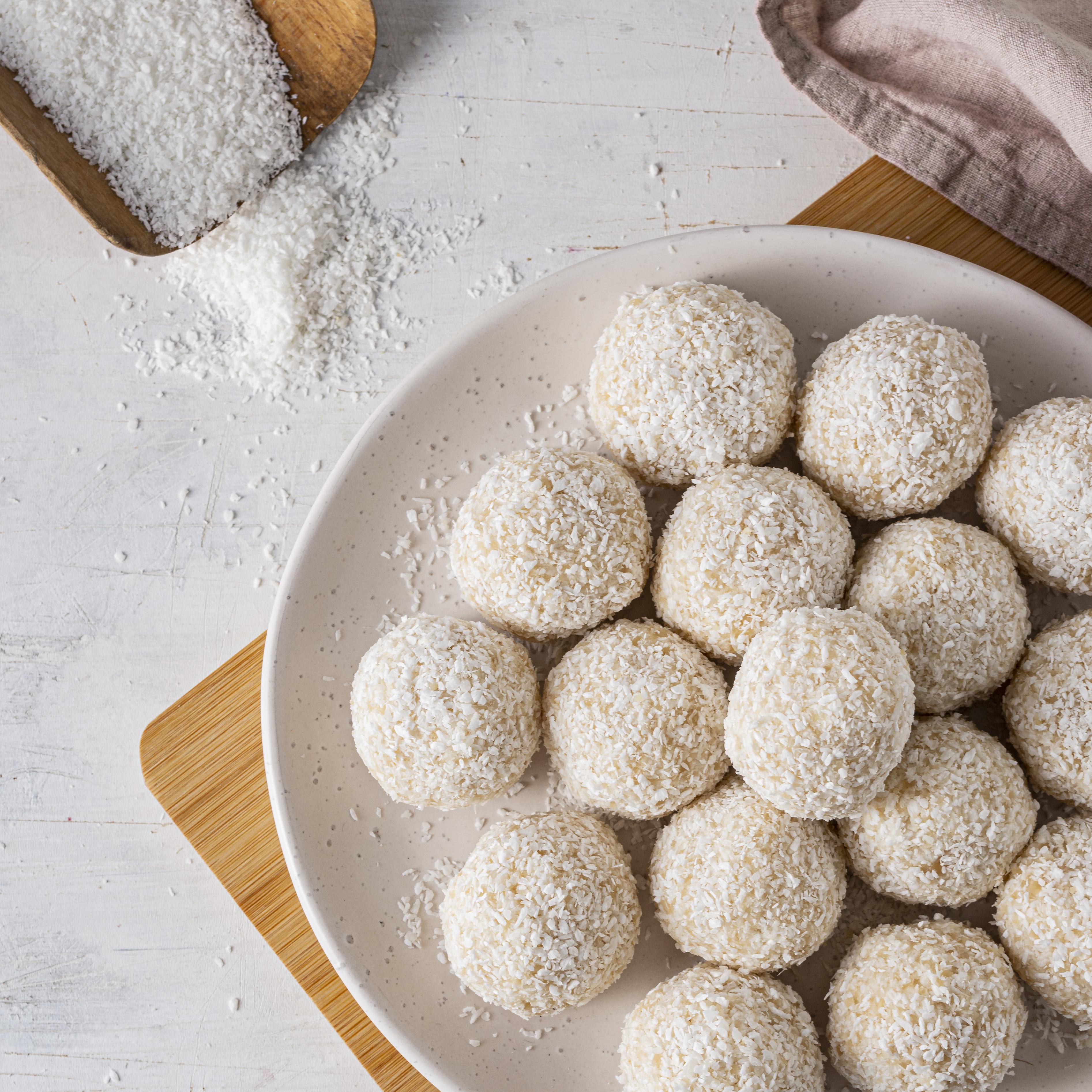 Prato com v&aacute;rias bolinhas de coco cobertas de coco ralado, sobre uma mesa branca, ao lado de uma tigela com coco ralado