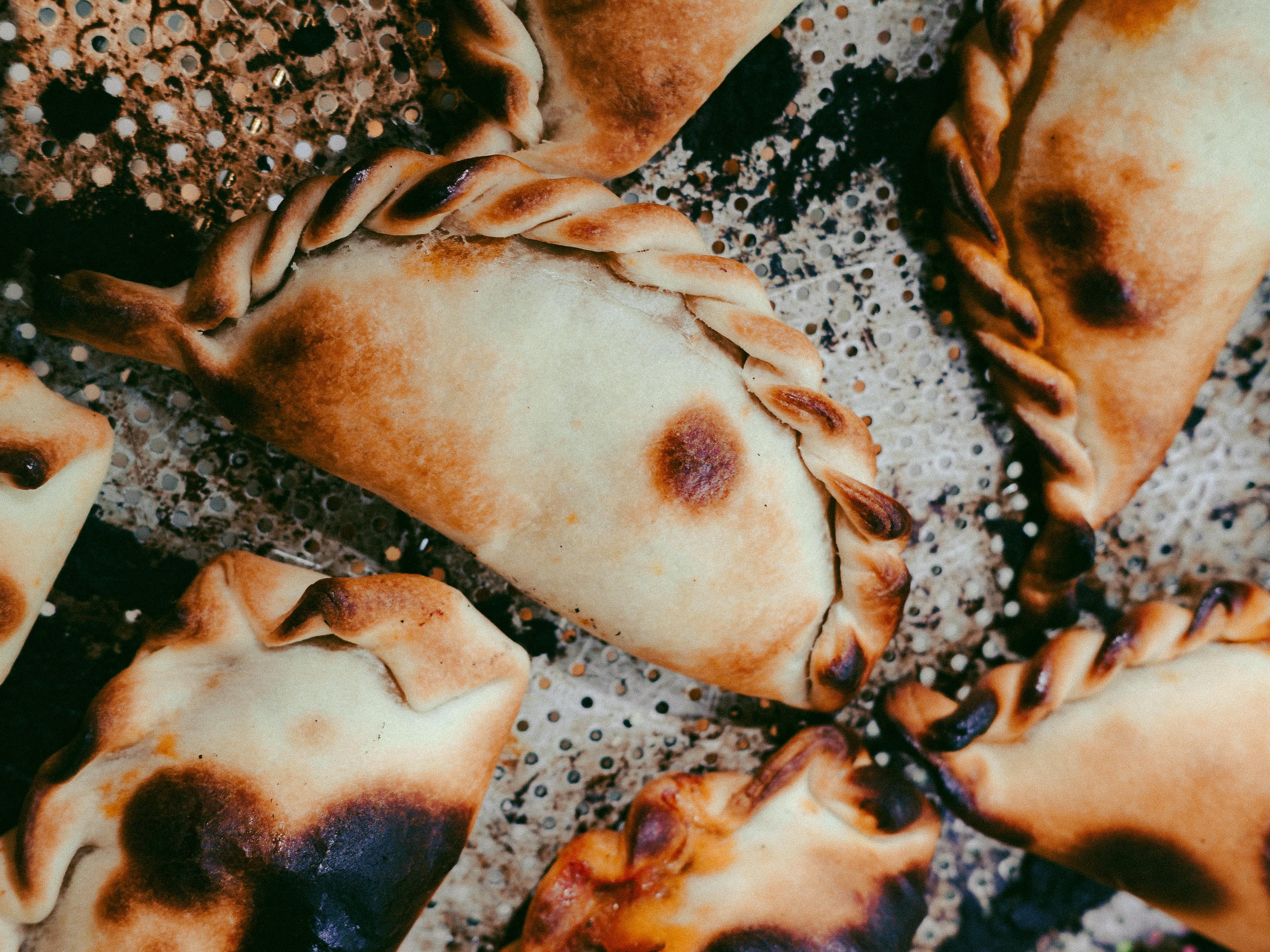 Empanadas assadas com massa dourada e bordas tran&ccedil;adas, algumas com pontos escuros de tostado, sobre uma assadeira met&aacute;lica perfurada