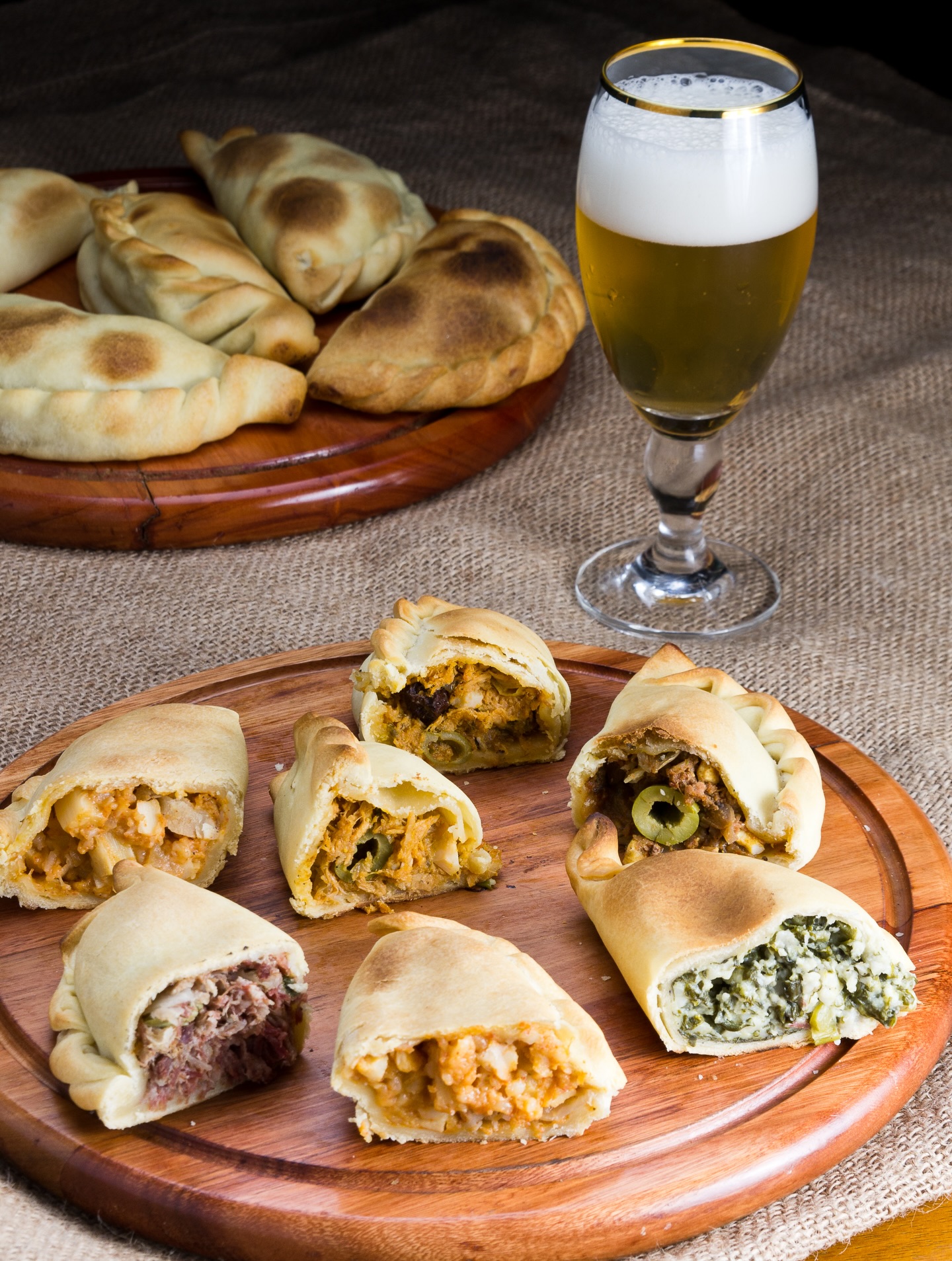 Empanadas assadas com recheios variados, algumas abertas mostrando carne, frango, queijo e espinafre, servidas em t&aacute;buas de madeira com um copo de cerveja clara ao lado