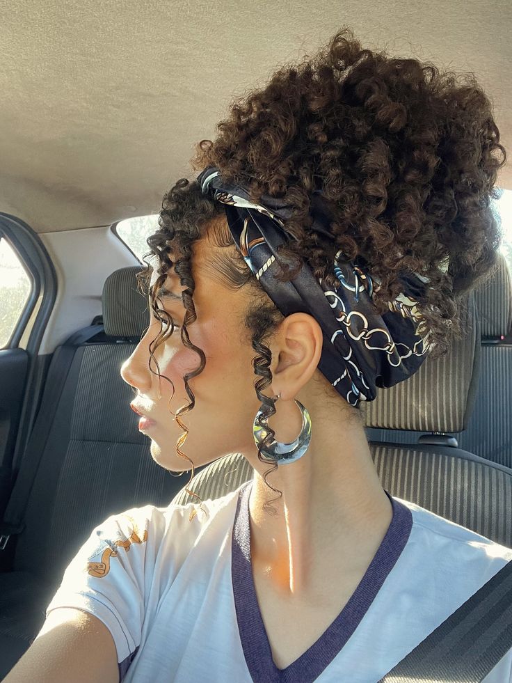 Mulher de perfil, com cabelo cacheado preso por um len&ccedil;o preto estampado, usando brincos de argola grandes e camiseta branca com gola V roxa, dentro de um carro