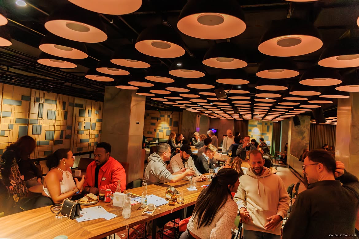 Pessoas conversam em um restaurante ou bar com ilumina&ccedil;&atilde;o ambiente. O teto &eacute; coberto por lumin&aacute;rias pendentes em formato de c&uacute;pula, emitindo uma luz quente. V&aacute;rias mesas de madeira est&atilde;o ocupadas por grupos de pessoas, algumas comendo e bebendo, outras engajadas em conversas animadas. As paredes t&ecirc;m um padr&atilde;o geom&eacute;trico em tons de bege e cinza. A atmosfera &eacute; descontra&iacute;da e social