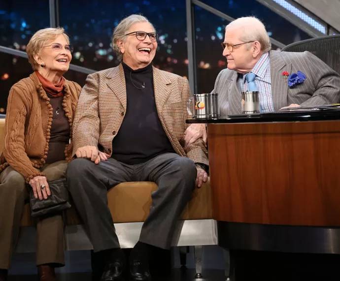 Tr&ecirc;s pessoas idosas em um programa de TV. Uma mulher de &oacute;culos e casaco marrom sorri ao lado de um homem de &oacute;culos, blazer xadrez e gola alta preta, que ri abertamente. &Agrave; direita, outro homem de terno risca de giz, camisa listrada e gravata azul, olha para eles com um sorriso discreto. H&aacute; canecas sobre a mesa.