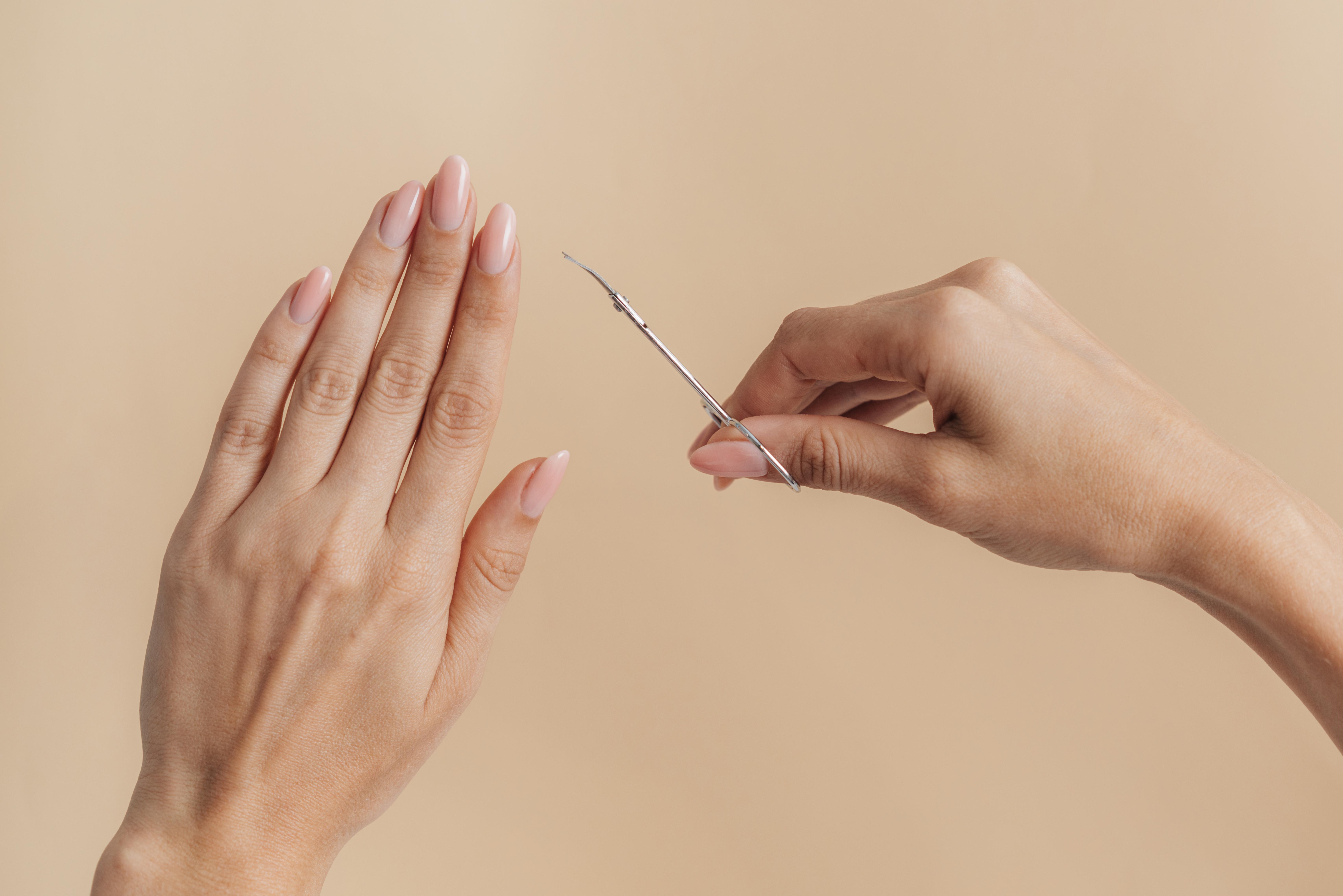 Duas m&atilde;os femininas em fundo bege claro. A m&atilde;o esquerda est&aacute; aberta, com unhas longas e rosadas. A m&atilde;o direita segura uma tesoura de cut&iacute;cula prateada, apontando para a m&atilde;o esquerda