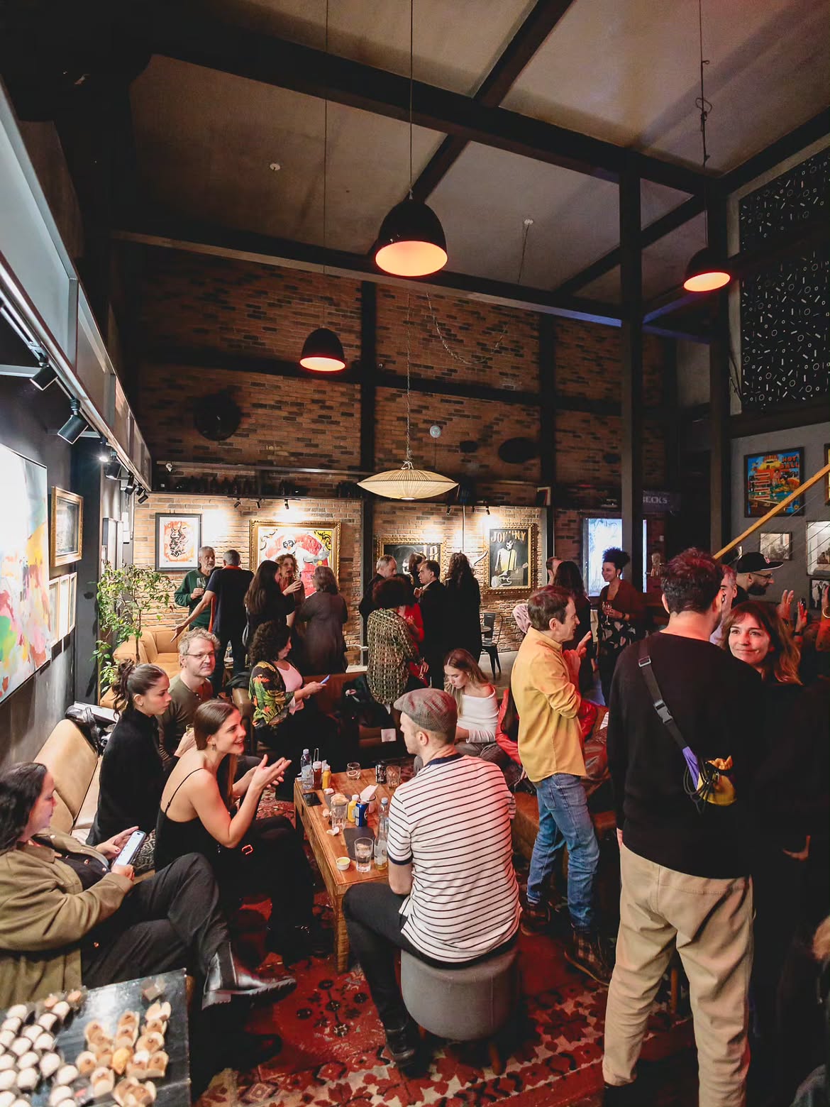 Pessoas conversam em um bar ou galeria de arte com paredes de tijolos, quadros coloridos e ilumina&ccedil;&atilde;o baixa. Alguns est&atilde;o sentados em sof&aacute;s e bancos, outros em p&eacute;, socializando. O ambiente &eacute; descontra&iacute;do e animado, com um tapete persa no ch&atilde;o.