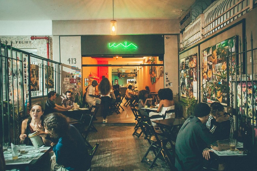 Pessoas sentadas em mesas de um bar com ilumina&ccedil;&atilde;o baixa e neon verde escrito KAIA na entrada. Mulheres e homens conversam e bebem em um ambiente descontra&iacute;do, com paredes decoradas por colagens e grades de ferro. Uma l&acirc;mpada pendente ilumina o centro do local