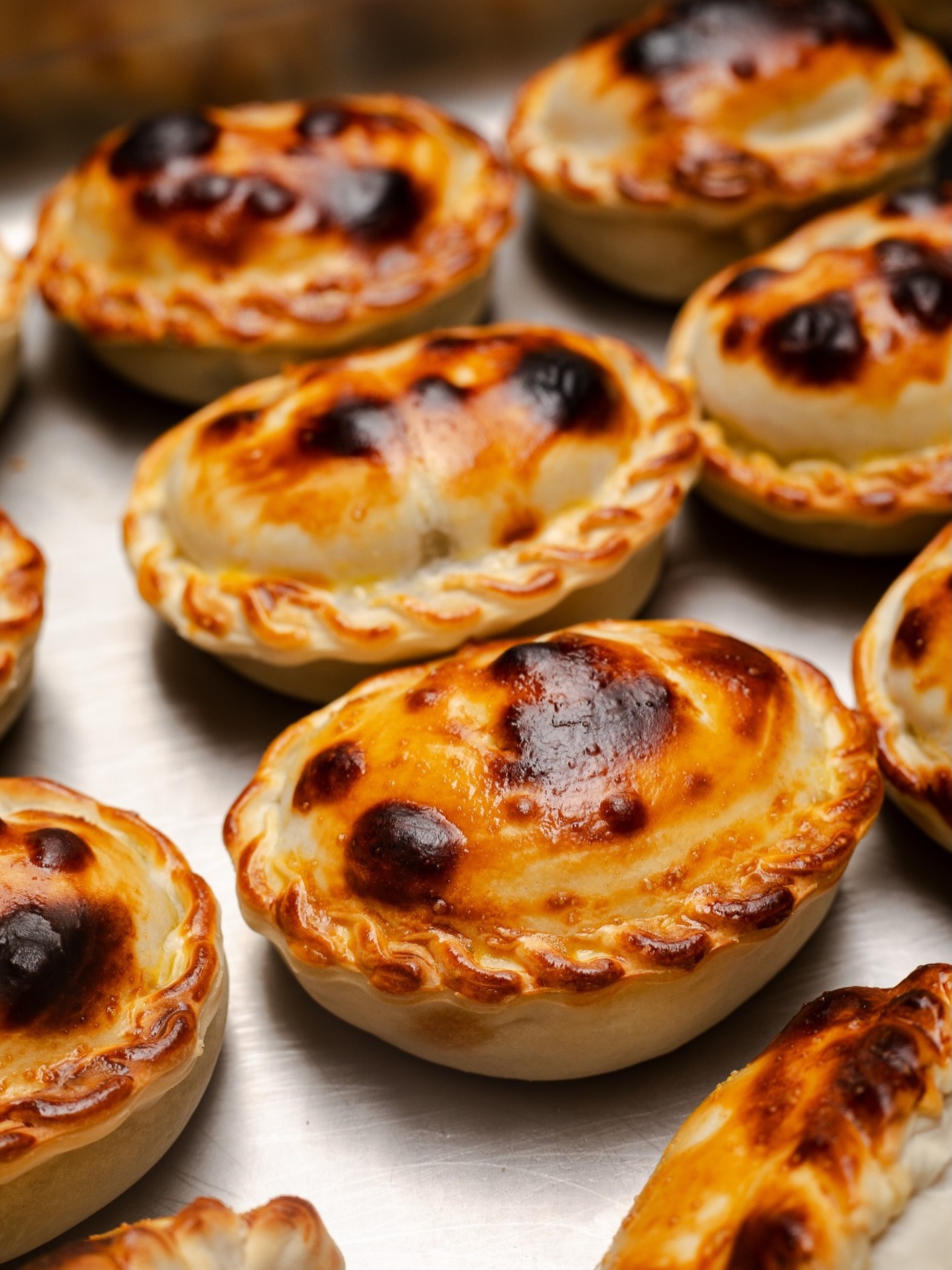 Muitas empanadas assadas, com massa dourada e partes mais escuras, dispostas em uma assadeira met&aacute;lica