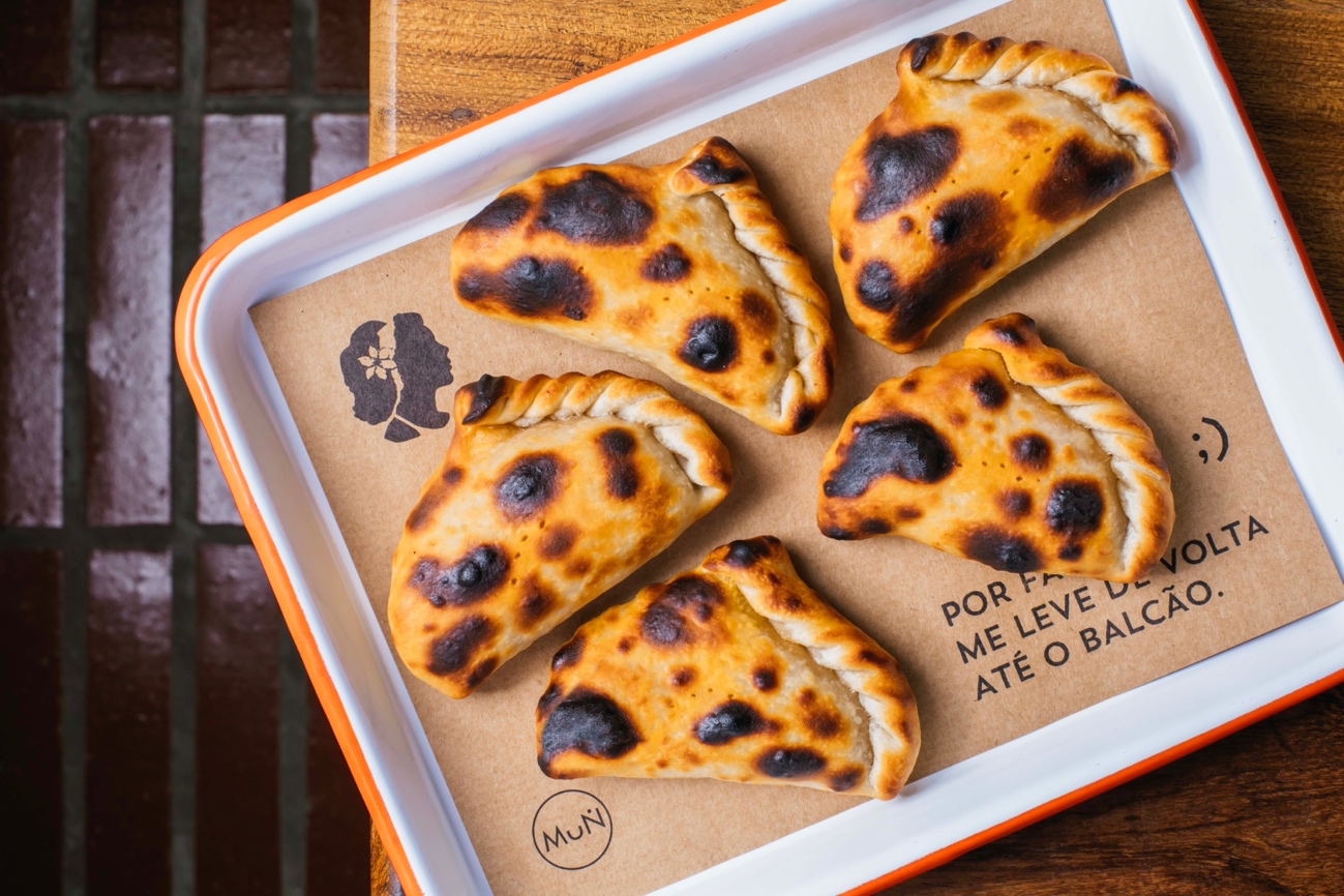 Seis empanadas assadas com manchas escuras de forno, servidas em uma bandeja branca com borda laranja sobre papel pardo. O papel tem um logo de duas silhuetas de perfil e a frase POR FAVOR ME LEVE DE VOLTA AT&Eacute; O BALC&Atilde;O