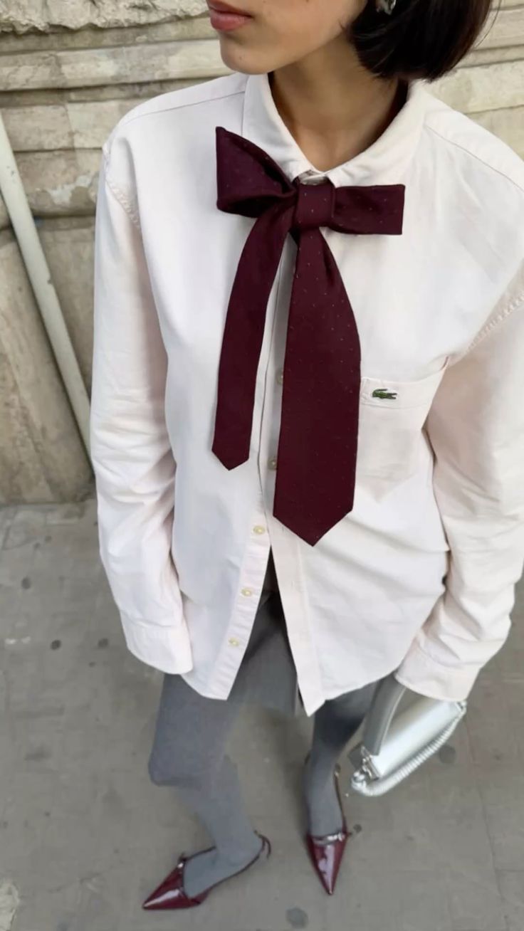 Pessoa de pele clara, cabelo curto escuro, vestindo camisa social branca com gravata borboleta vinho, cal&ccedil;a cinza e sapatos de bico fino vinho, segurando uma bolsa branca. O fundo &eacute; uma parede de pedra e ch&atilde;o de cimento