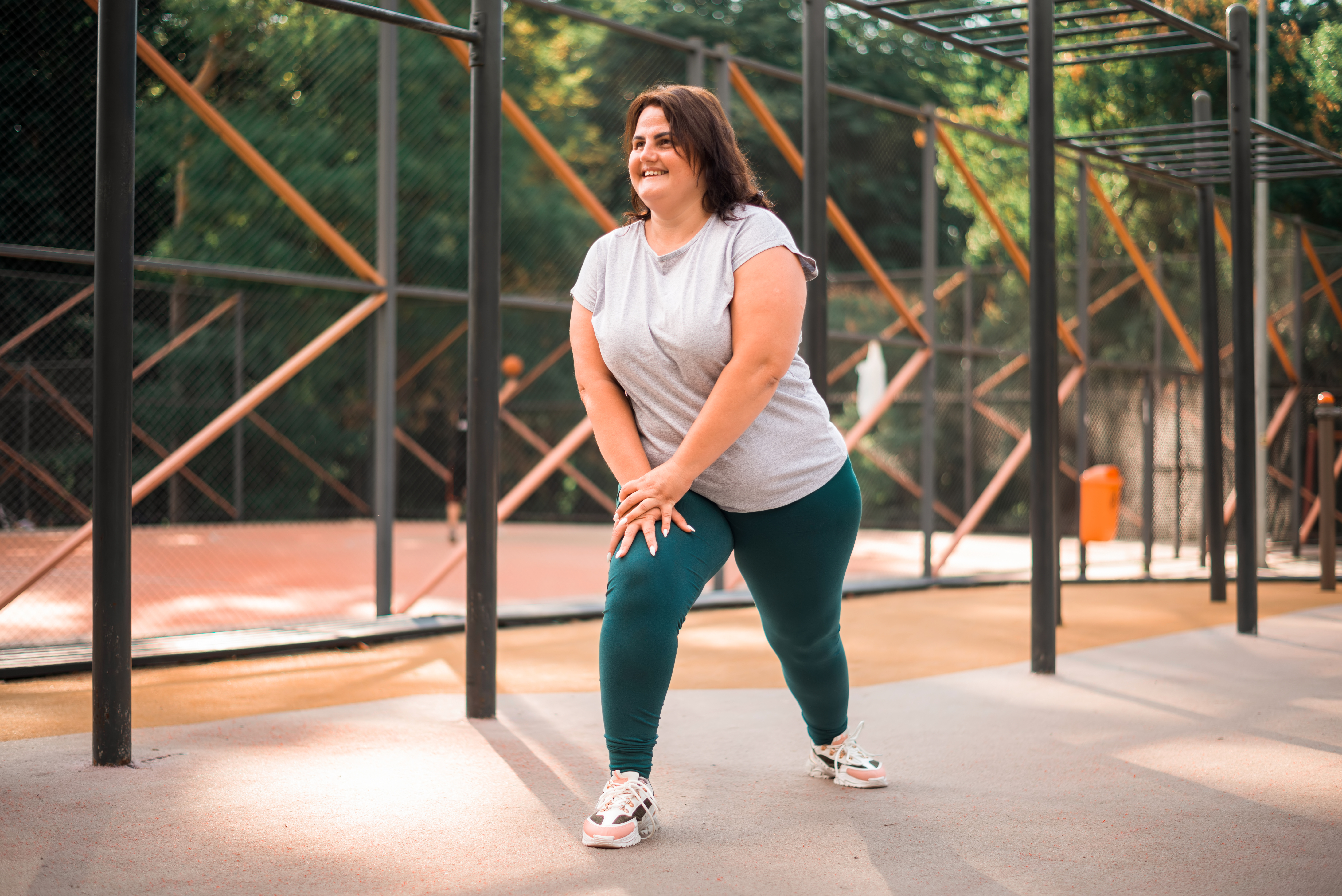 Mulher sorridente de cabelos castanhos, camiseta cinza e legging verde-escura, alongando-se com as m&atilde;os nos joelhos em uma quadra esportiva ao ar livre