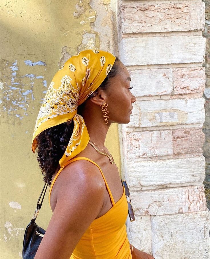 Mulher de pele escura, perfil, usando bandana amarela estampada, top amarelo de al&ccedil;as finas, colar dourado e brincos dourados com a palavra amor em escrita cursiva. Ela est&aacute; em frente a uma parede de tijolos amarelados e brancos, com uma bolsa preta no ombro esquerdo