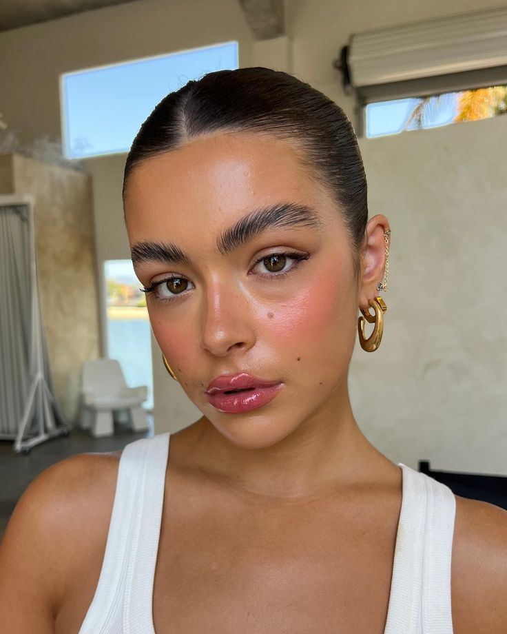 Mulher jovem com pele bronzeada, cabelo escuro preso e maquiagem natural, incluindo blush rosado e gloss nos l&aacute;bios. Ela usa um top branco de al&ccedil;as e brincos dourados grandes, com um piercing de corrente na orelha esquerda. O fundo &eacute; desfocado, com tons claros e uma cadeira branca vis&iacute;vel