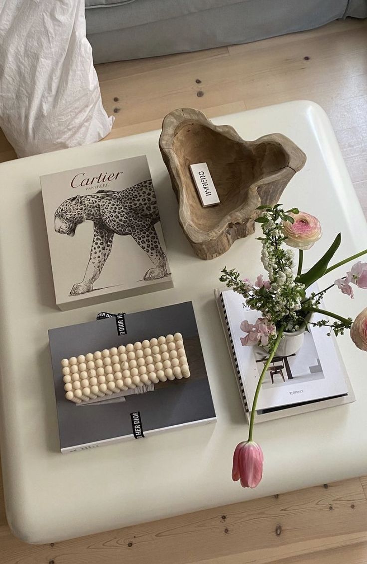 Uma mesa de centro branca com tr&ecirc;s livros de capa dura, um vaso de flores rosa e brancas, e uma tigela de madeira com uma barra de sabonete Byredo. O livro superior exibe um leopardo preto e branco com o t&iacute;tulo Cartier Panth&egrave;re. Um livro cinza com Dior na lombada tem um objeto bege de bolinhas em cima. Um livro aberto com uma imagem de cadeira e o vaso de flores. Uma tulipa rosa pende da mesa