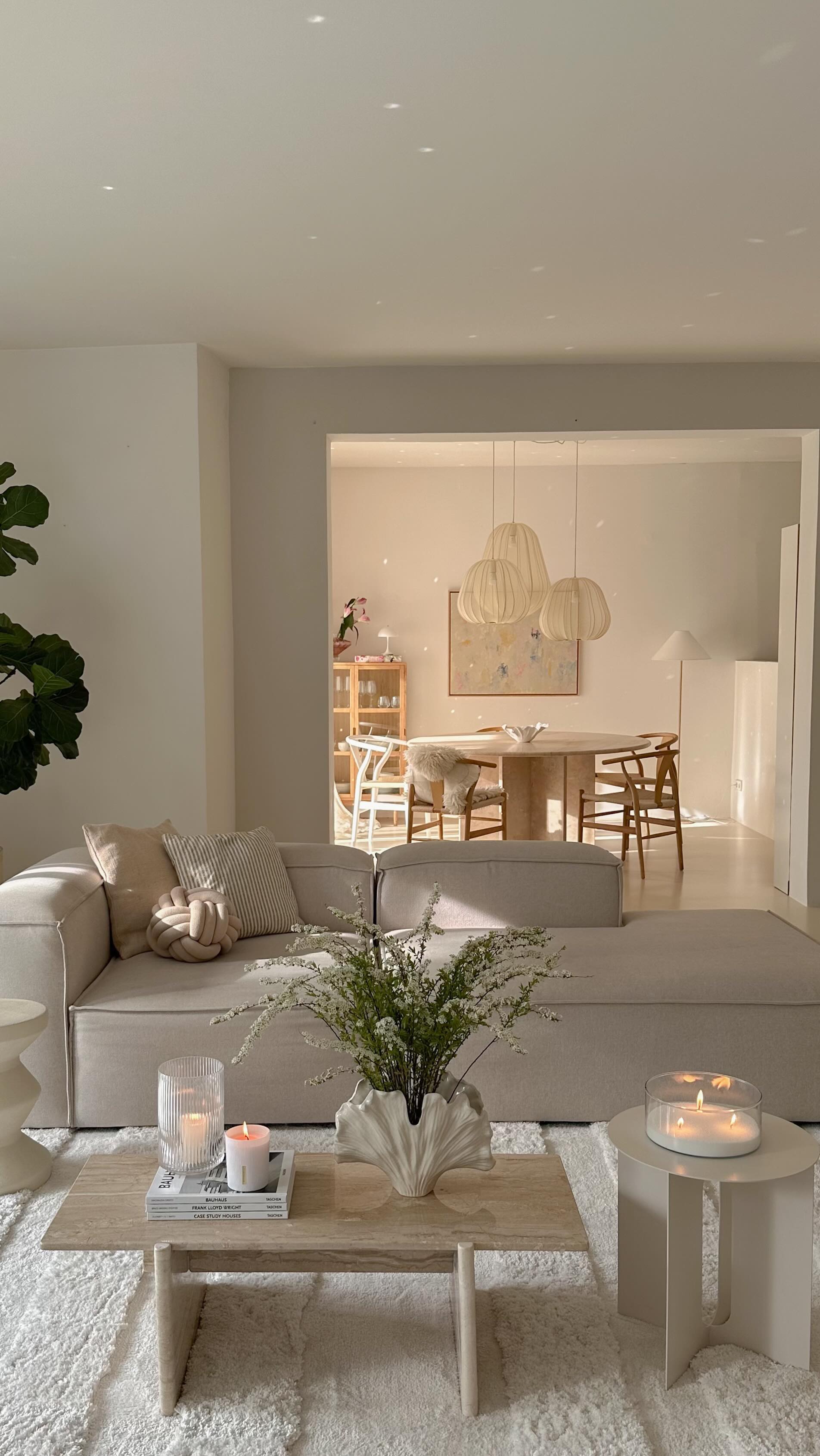 Sala de estar e jantar em tons claros, com sof&aacute; bege, almofadas decorativas e mesa de centro de m&aacute;rmore com livros, velas e um vaso de flores brancas. Ao fundo, sala de jantar com mesa redonda, cadeiras de madeira e lumin&aacute;rias pendentes, tudo sob luz natural