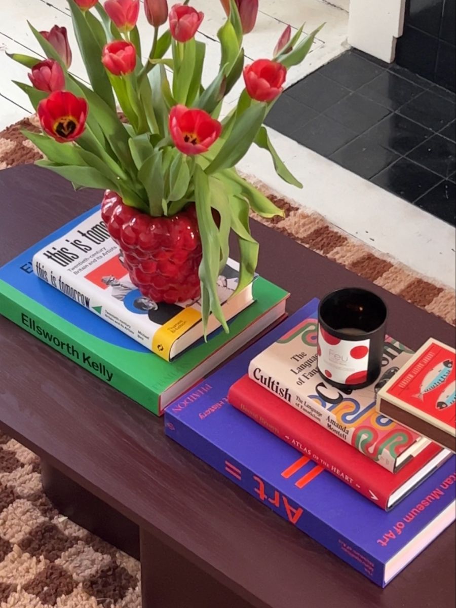 Vaso vermelho texturizado com tulipas vermelhas vibrantes, sobre uma pilha de livros coloridos em uma mesa de centro de madeira escura. Ao lado, uma vela preta e uma caixa de f&oacute;sforos decorada. Um tapete marrom e bege aparece no ch&atilde;o