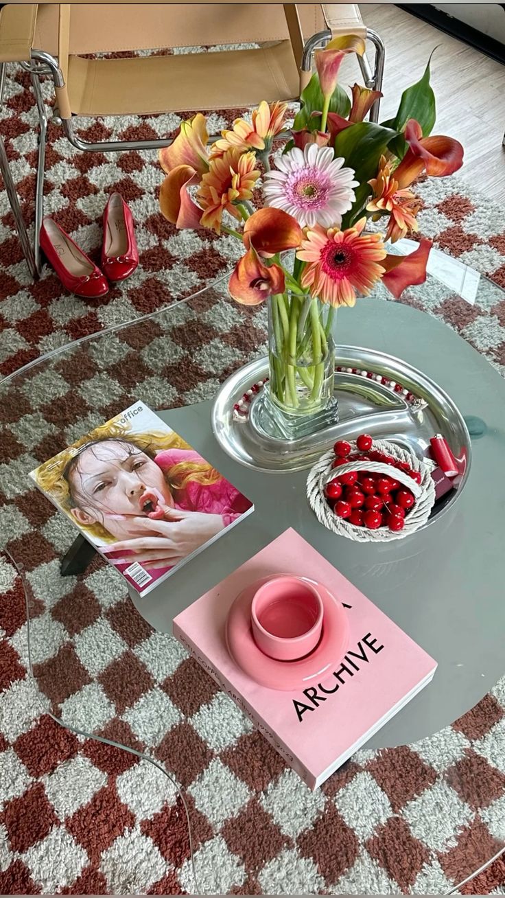 Mesa de centro de vidro com arranjo de flores coloridas, revista Office, livro rosa Archive com x&iacute;cara rosa em cima, e cesta de cerejas artificiais. No ch&atilde;o, um tapete xadrez marrom e branco com sapatilhas vermelhas.