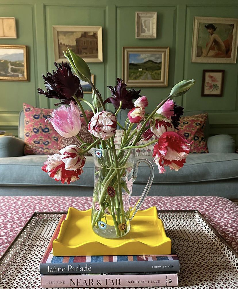 Um jarro de vidro com tulipas coloridas, incluindo tons de rosa, vermelho, branco e roxo escuro, sobre uma bandeja amarela e livros, em uma mesa de centro com estampa vermelha. Ao fundo, uma parede verde com quadros e um sof&aacute; azul com almofadas estampadas