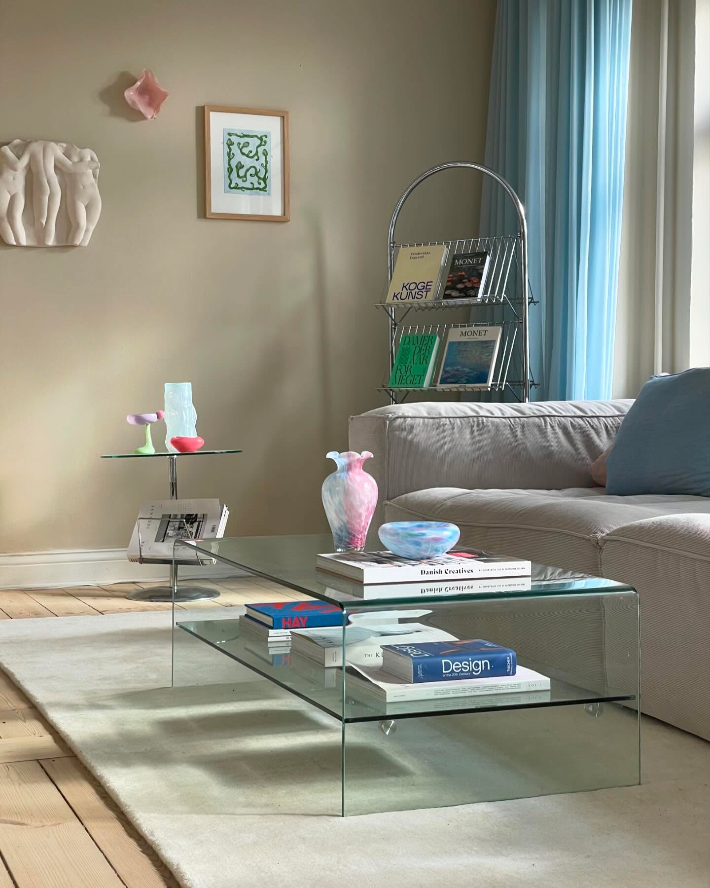 Sala de estar com sof&aacute; cinza, cortinas azuis e paredes bege. Uma mesa de centro de vidro exibe livros e vasos coloridos. Na parede, uma escultura branca e um quadro verde. Um revisteiro met&aacute;lico com revistas Monet e Koge Kunst fica ao lado do sof&aacute;. O ambiente tem piso de madeira e um tapete claro.