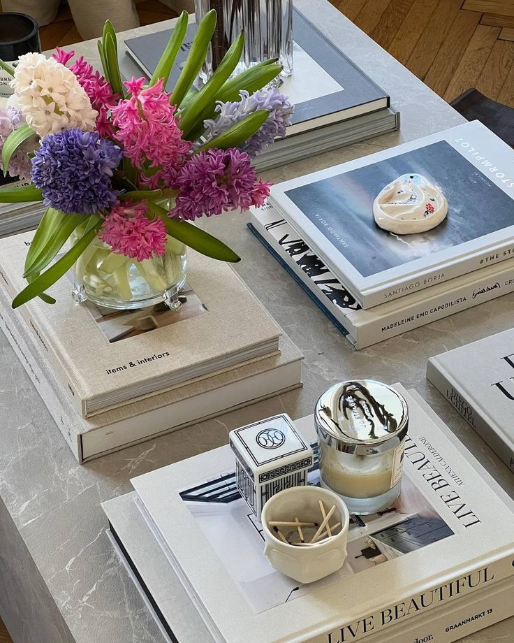 Arranjo de flores jacinto em tons de rosa, roxo e branco em um vaso de vidro, sobre uma pilha de livros de capa dura em uma mesa de m&aacute;rmore cinza. Ao lado, uma vela arom&aacute;tica e uma caixa de f&oacute;sforos decorativa.