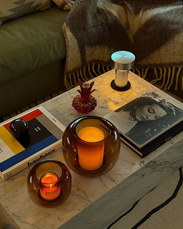 Mesa de m&aacute;rmore com dois casti&ccedil;ais de vidro &acirc;mbar e velas acesas, um livro Bauhaus e outro Rostros del Cine Mexicano, uma lumin&aacute;ria prateada e um objeto decorativo vermelho em formato de abacaxi. Ao fundo, um sof&aacute; verde com manta marrom