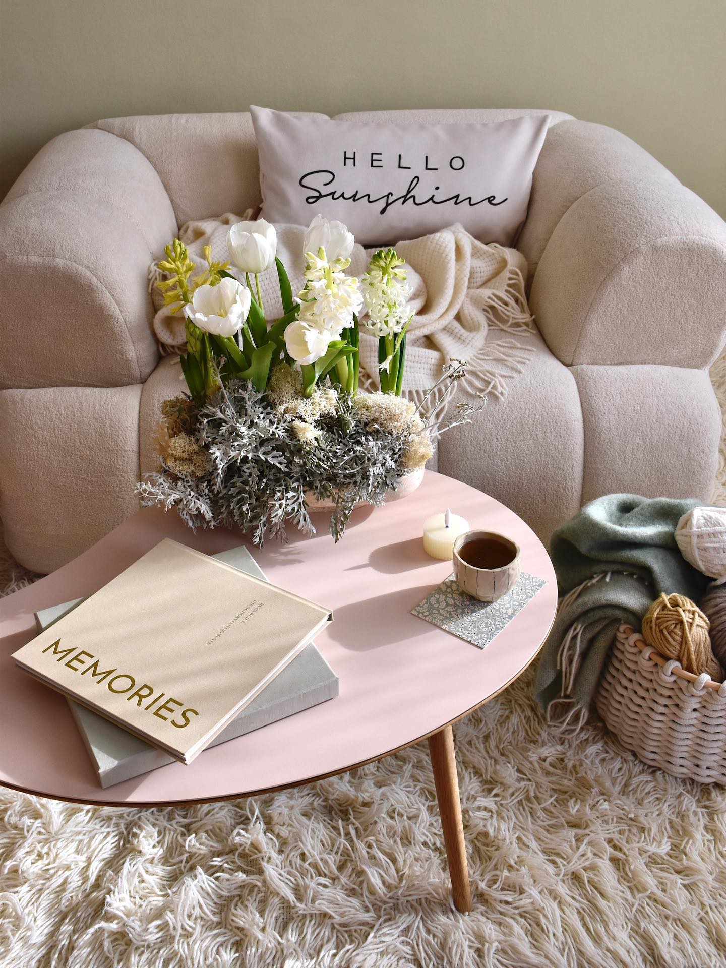 Ambiente aconchegante com poltrona creme, manta e almofada Hello Sunshine. Uma mesa de centro rosa tem um vaso de flores brancas, livros, uma vela e uma x&iacute;cara de ch&aacute;. No ch&atilde;o, um tapete felpudo e uma cesta com novelos de l&atilde;