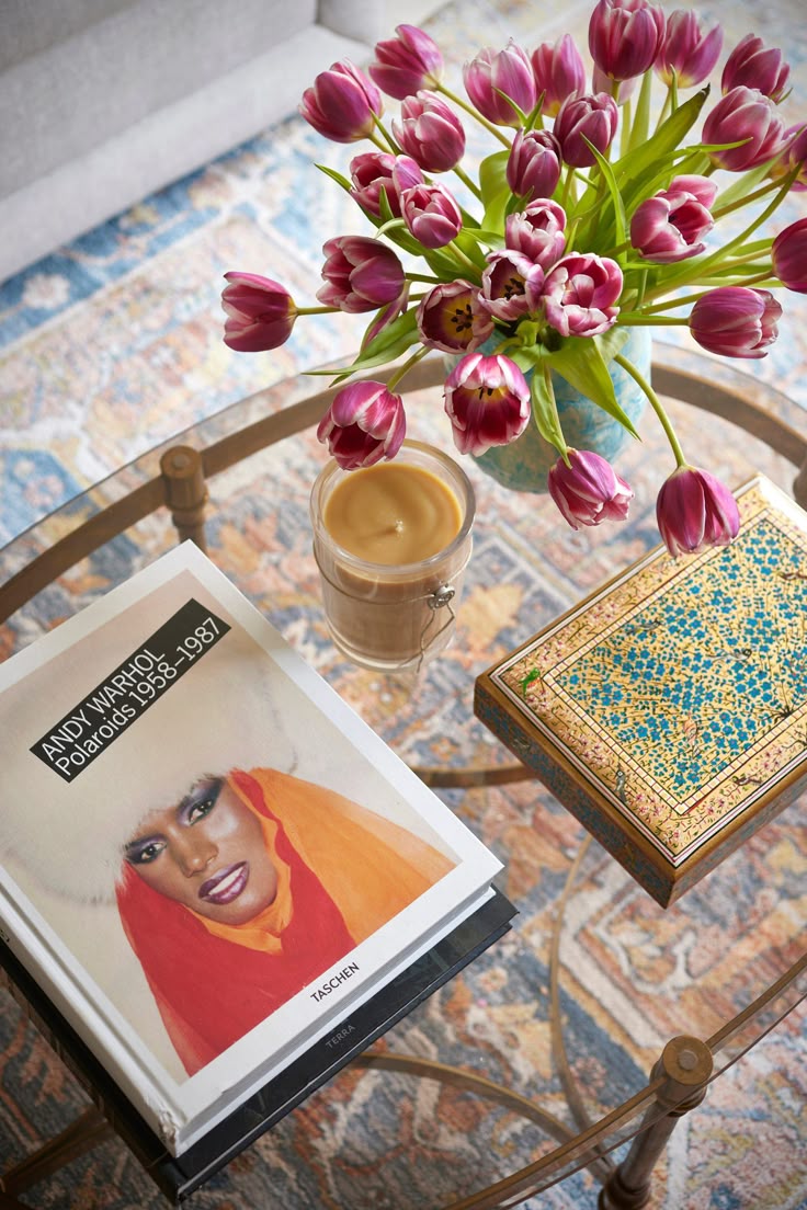 Mesa de centro com tampo de vidro, exibindo um vaso azul com tulipas roxas e brancas, uma vela acesa em copo de vidro, um livro Andy Warhol Polaroids 1958-1987 com a capa de Grace Jones, e uma caixa decorada com padr&otilde;es azuis e dourados. O tapete estampado em tons de azul e bege complementa o ambiente