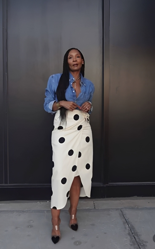 Mulher negra de cabelos longos e lisos, vestindo camisa jeans azul e saia envelope branca com bolinhas pretas, posa em frente a uma parede escura. Ela usa um colar dourado, pulseiras e sapatos de salto com bico em formato de cora&ccedil;&atilde;o.