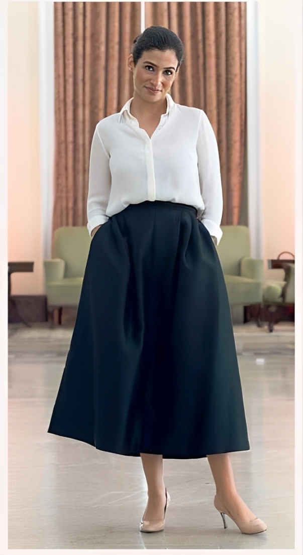 Mulher de pele morena, cabelo escuro preso, sorri levemente para a c&acirc;mera. Veste camisa social branca e saia midi preta com pregas, m&atilde;os nos bolsos. Cal&ccedil;a scarpin nude de salto fino. Ao fundo, cortinas marrons e poltronas verdes emba&ccedil;adas