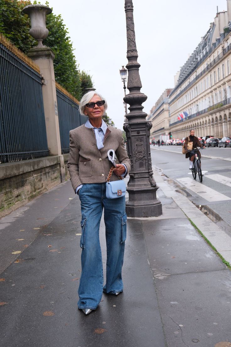 Mulher idosa de cabelos grisalhos curtos, &oacute;culos escuros, blazer xadrez marrom, camisa azul clara, cal&ccedil;a jeans cargo e sapatos prateados, segurando uma bolsa azul clara e um jornal enrolado, caminhando em uma cal&ccedil;ada molhada com folhas secas. Ao fundo, um poste de luz ornamentado, um muro com port&atilde;o e pr&eacute;dios antigos. Uma pessoa de bicicleta passa na rua.