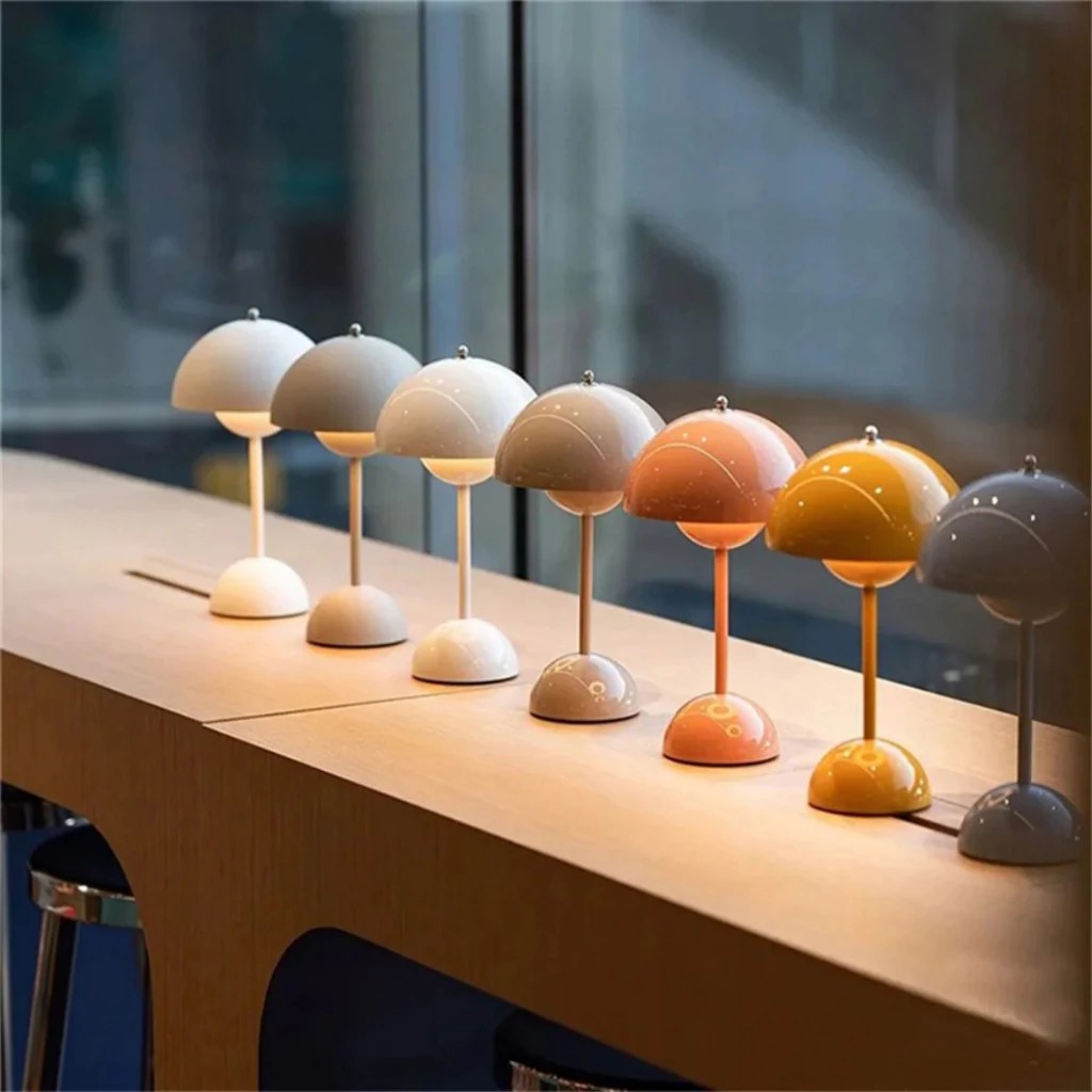 Sete lumin&aacute;rias de mesa em formato de cogumelo, com c&uacute;pulas e bases em tons pastel variados, alinhadas sobre um balc&atilde;o de madeira claro, iluminando suavemente o ambiente