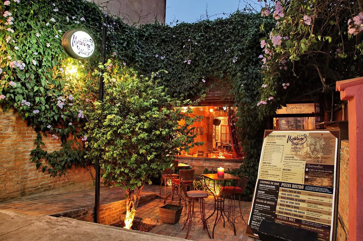 Fachada de um bistr&ocirc; &agrave; noite, com paredes de tijolos cobertas por hera e flores lilases. Uma placa redonda iluminada com o nome Madeleine se destaca. Em frente, um pequeno p&aacute;tio com mesas e cadeiras de ferro, uma vela acesa sobre uma mesa, e um card&aacute;pio grande em um cavalete. Um arbusto verde no centro do p&aacute;tio &eacute; iluminado por baixo, criando um ambiente acolhedor. A entrada do bistr&ocirc;, com luzes quentes, &eacute; vis&iacute;vel ao fundo