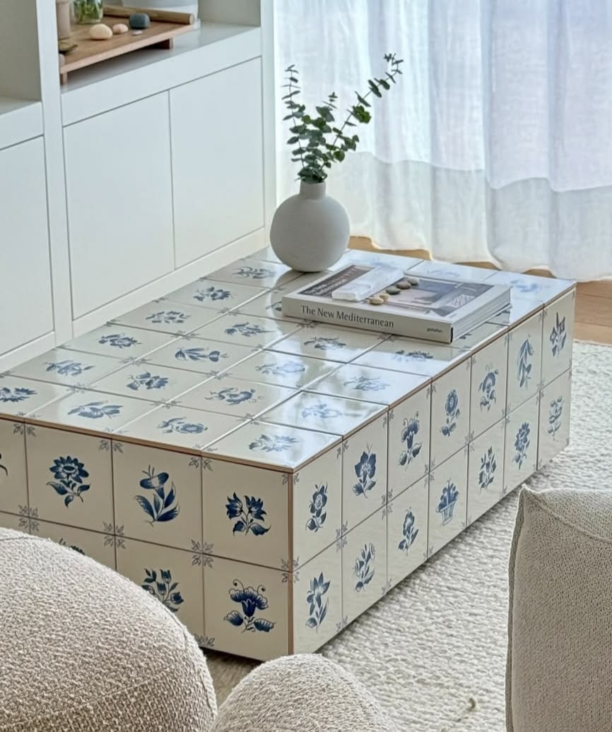Mesa de centro retangular com azulejos brancos e desenhos florais azuis, um vaso branco com folhagens verdes e um livro The New Mediterranean sobre ela. Ao fundo, arm&aacute;rios brancos e uma cortina transl&uacute;cida. Em primeiro plano, parte de um sof&aacute; claro.