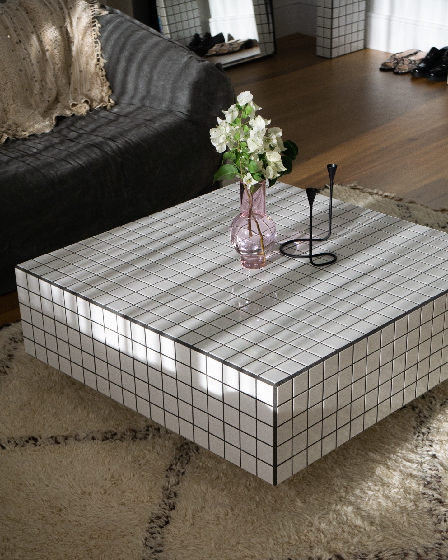 Mesa de centro quadrada com azulejos brancos e rejunte preto, sobre um tapete felpudo bege. Um vaso rosa com flores brancas e um casti&ccedil;al preto est&atilde;o sobre a mesa. Ao fundo, um sof&aacute; cinza e um espelho refletindo sapatos.