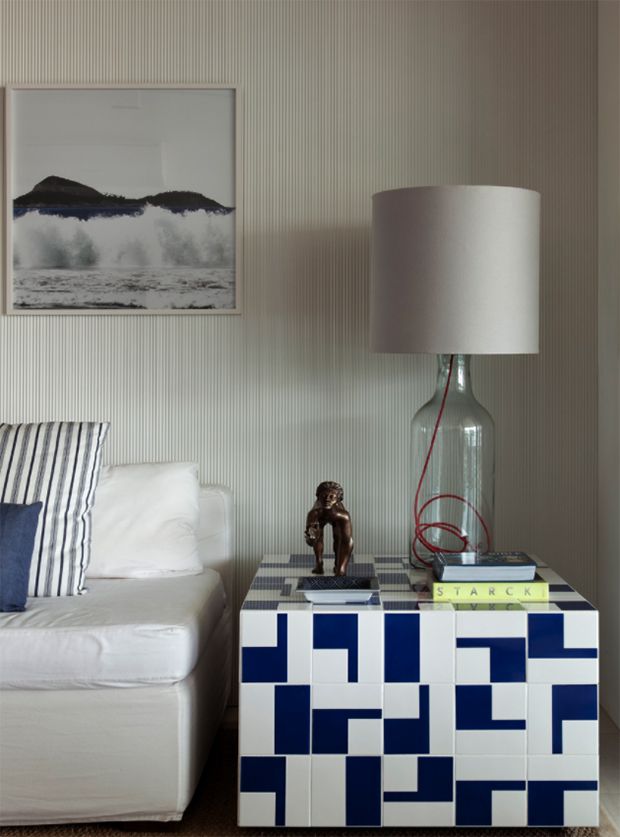 Quarto com parede listrada clara, quadro de ondas fortes, sof&aacute; branco com almofadas listradas e azuis, e mesa de cabeceira azul e branca com lumin&aacute;ria de garrafa de vidro e livro STARCK