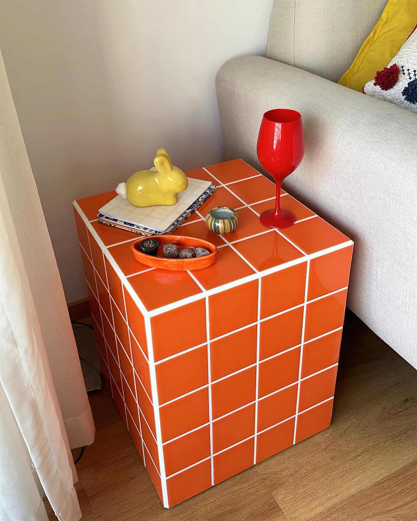 Mesa de cabeceira c&uacute;bica laranja com azulejos brancos, ao lado de um sof&aacute; bege. Sobre a mesa, um coelho de cer&acirc;mica amarelo, um copo de vinho vermelho, um prato com doces e um pequeno vaso listrado. Cortina branca &agrave; esquerda e piso de madeira.