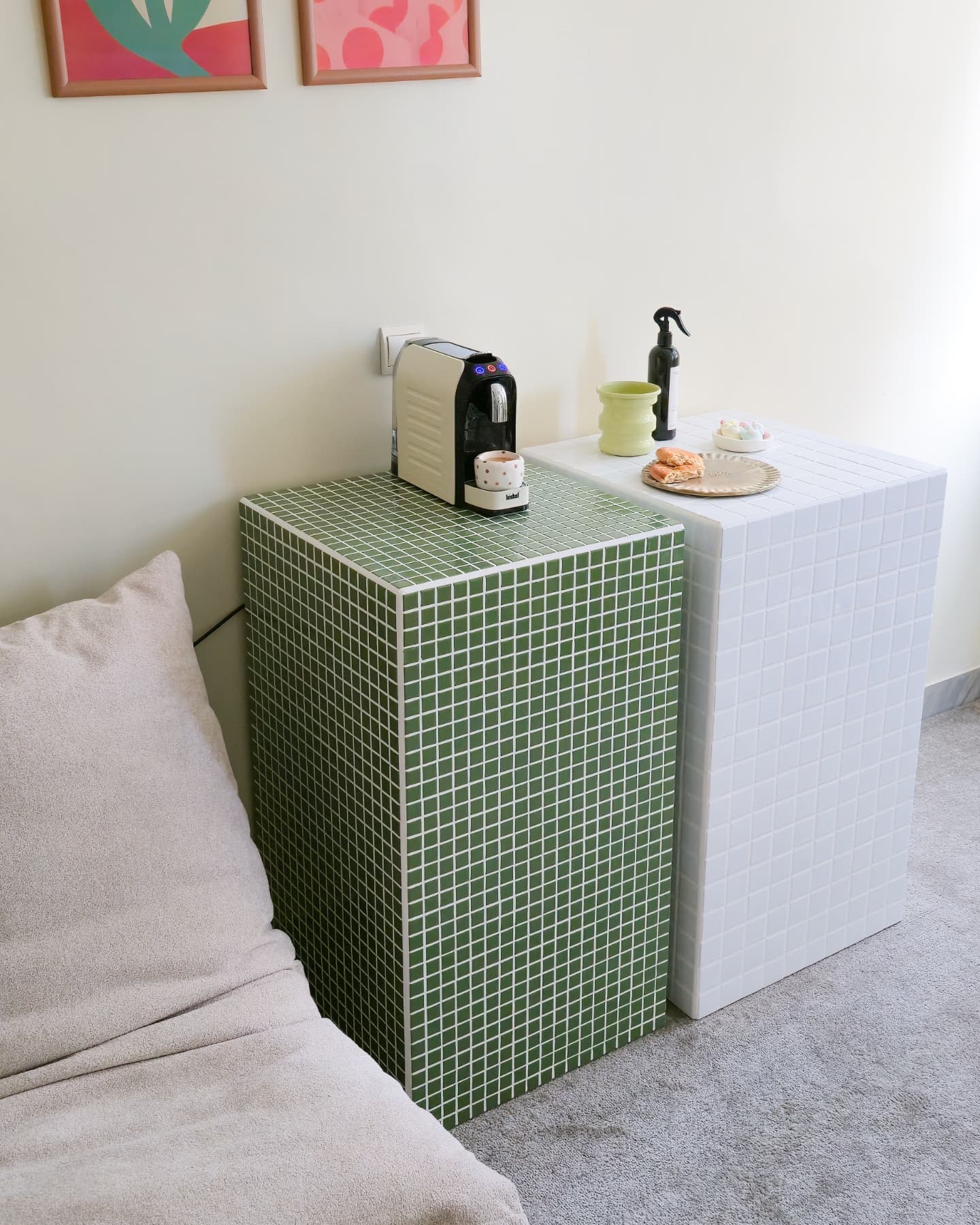 Duas mesas de cabeceira quadradas, uma verde e outra branca, com uma m&aacute;quina de caf&eacute; e x&iacute;cara na verde, e um prato com biscoitos na branca. Ao lado esquerdo, um sof&aacute; bege com almofada.