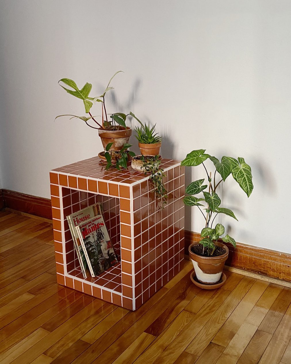 Mesa de cabeceira quadrada de azulejos marrons com rejunte branco, com tr&ecirc;s vasos de plantas no topo e dois livros dentro de um nicho. &Agrave; direita, um vaso de planta maior no ch&atilde;o de madeira, contra uma parede branca