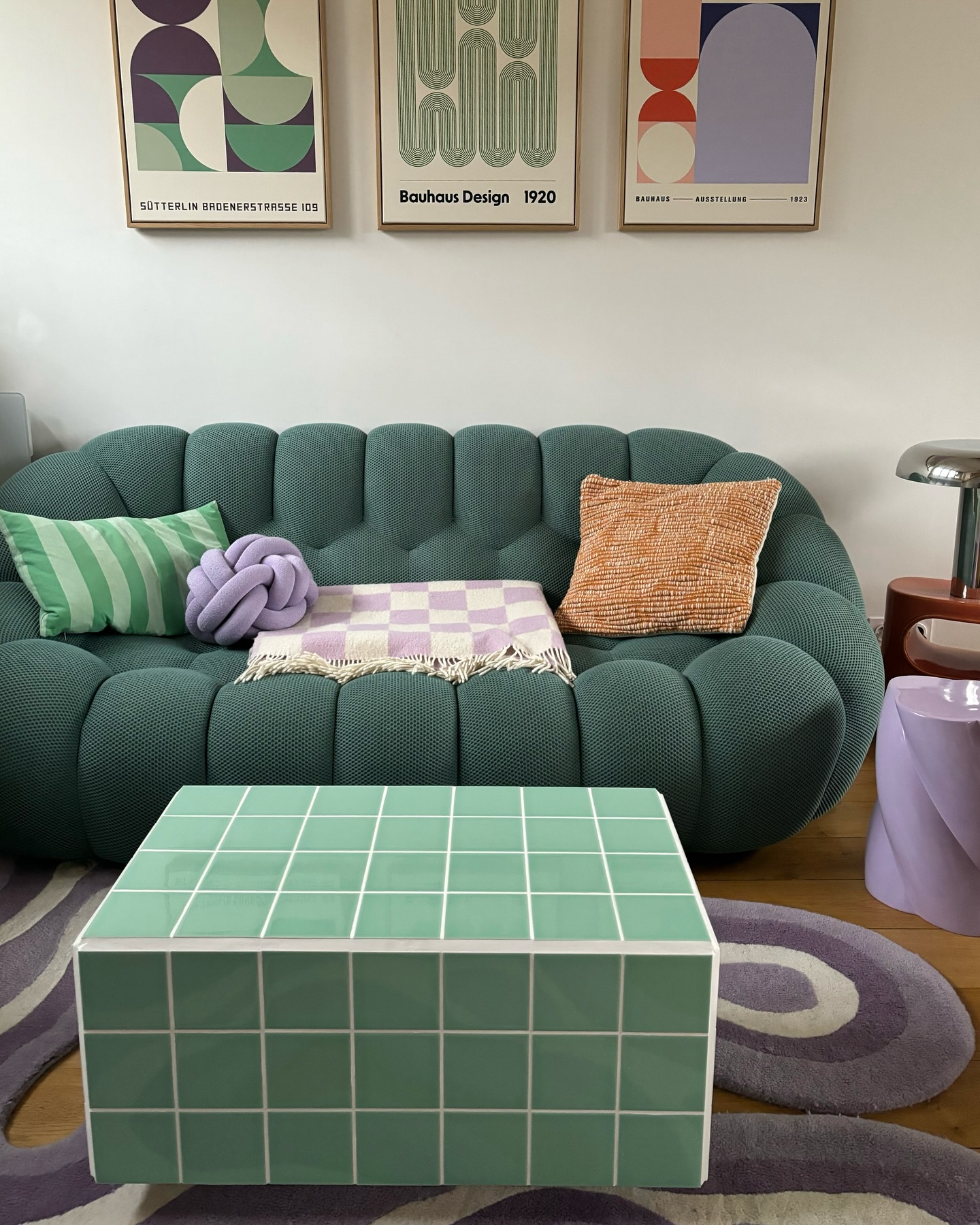 Sala de estar com sof&aacute; verde-escuro, almofadas listradas e lil&aacute;s, manta xadrez lil&aacute;s e branca. Uma mesa de centro verde-clara com azulejos, tapete roxo e quadros de arte Bauhaus na parede branca.