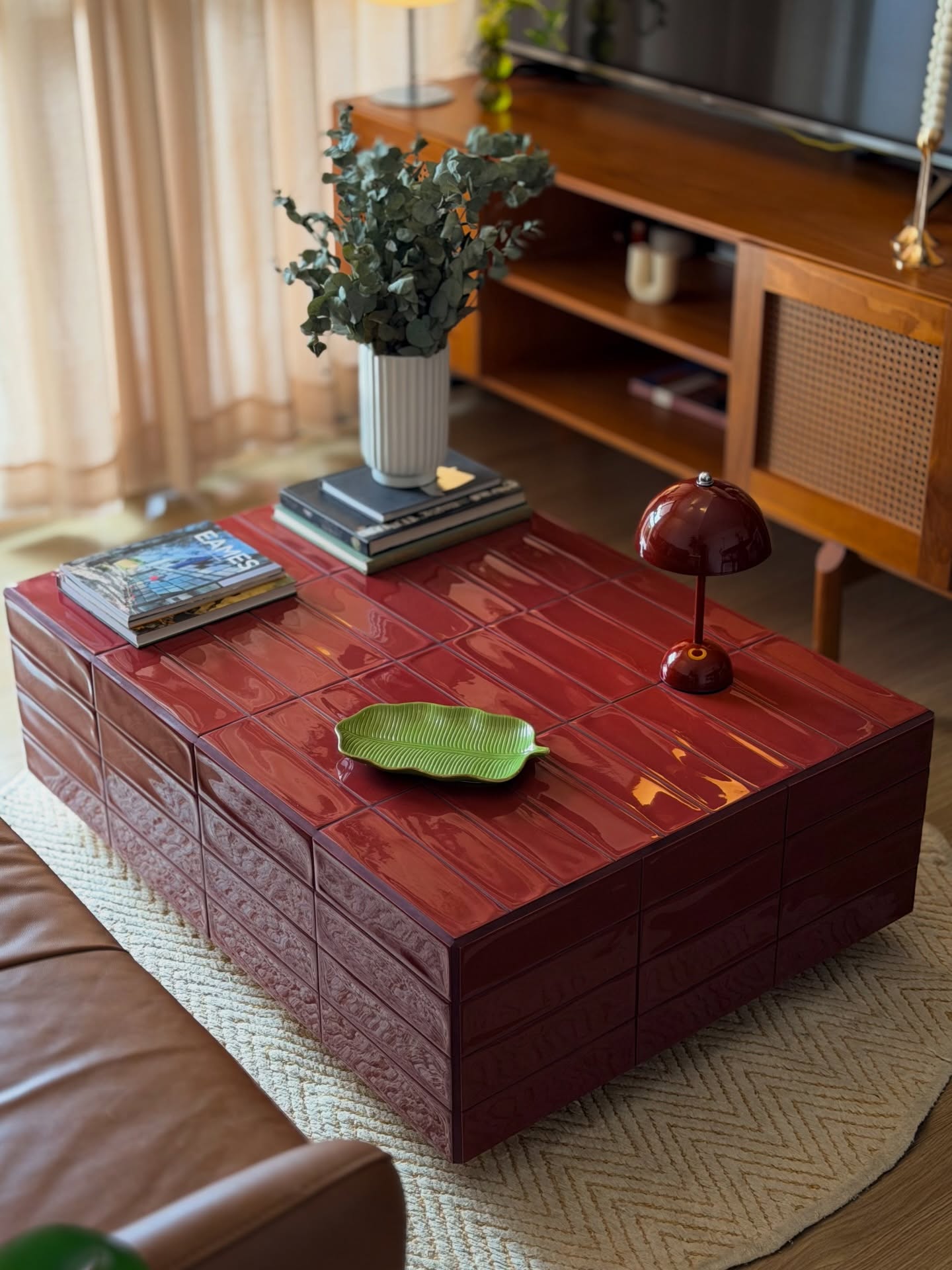 Mesa de centro retangular vermelha com acabamento em azulejos brilhantes, sobre um tapete claro de estampa geom&eacute;trica. Sobre a mesa, um vaso branco com folhagens verdes, uma lumin&aacute;ria pequena vermelha e uma travessa verde em formato de folha. Ao fundo, um m&oacute;vel de madeira com televis&atilde;o e um sof&aacute; de couro marrom