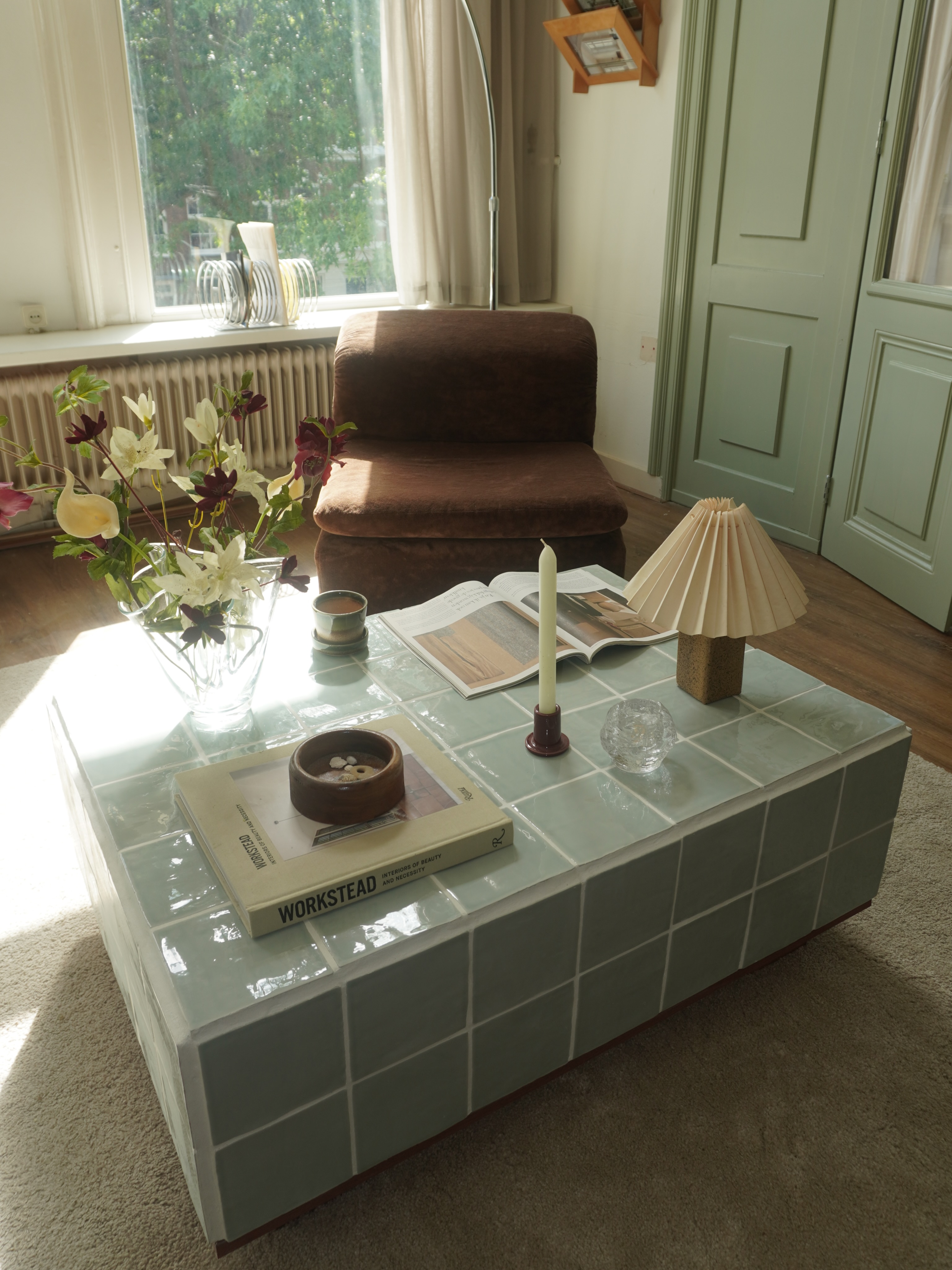 Sala de estar com luz solar entrando pela janela, iluminando uma mesa de centro retangular de azulejos verde-&aacute;gua. Sobre a mesa, um livro, uma vela acesa em casti&ccedil;al, uma lumin&aacute;ria plissada e um vaso de flores. Ao fundo, uma poltrona marrom e uma porta verde-clara