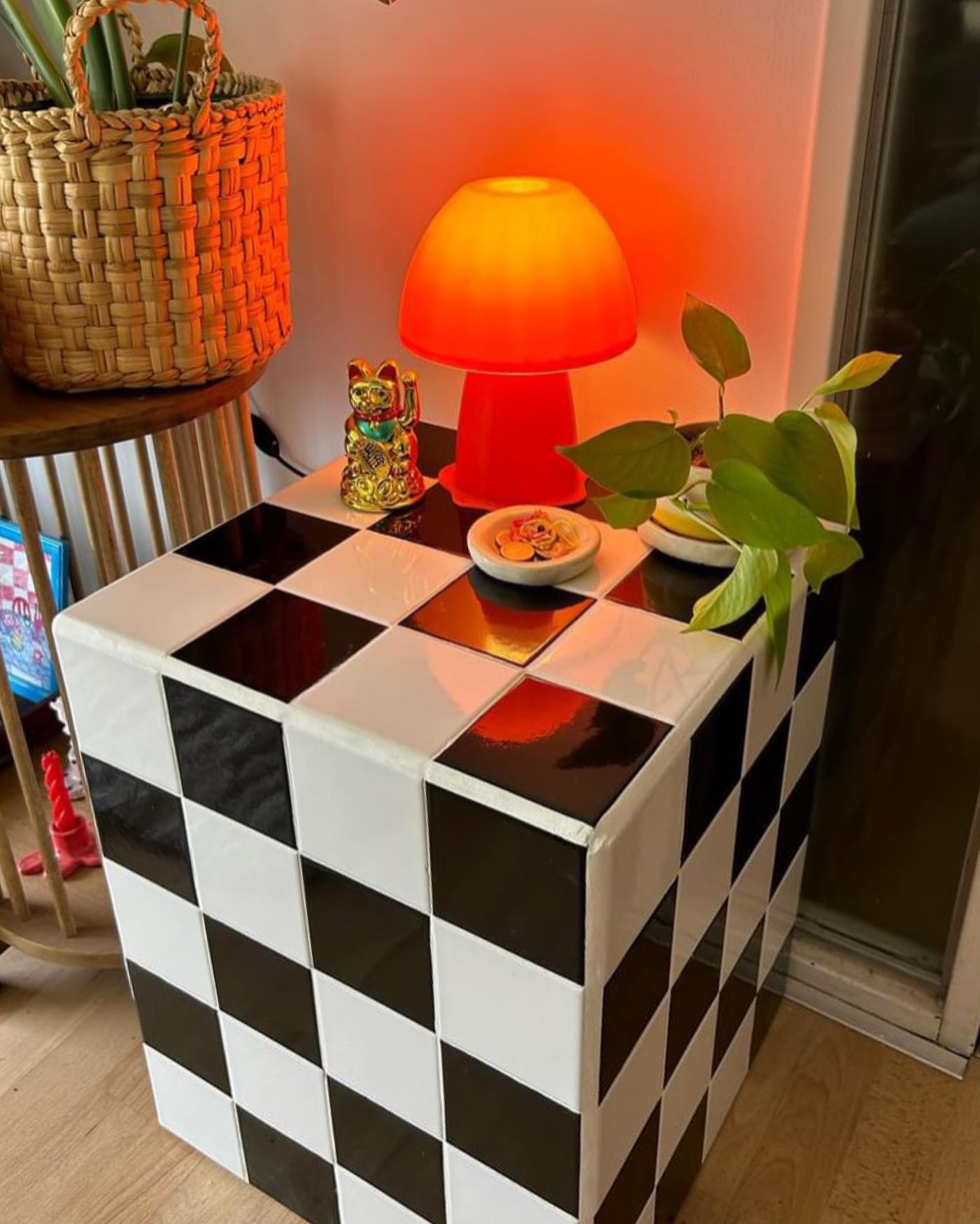 Mesa de cabeceira em formato de cubo com azulejos pretos e brancos, exibindo uma lumin&aacute;ria laranja em forma de cogumelo, um gato da sorte dourado, um pequeno pote com moedas e uma planta verde. Ao fundo, um cesto de palha.