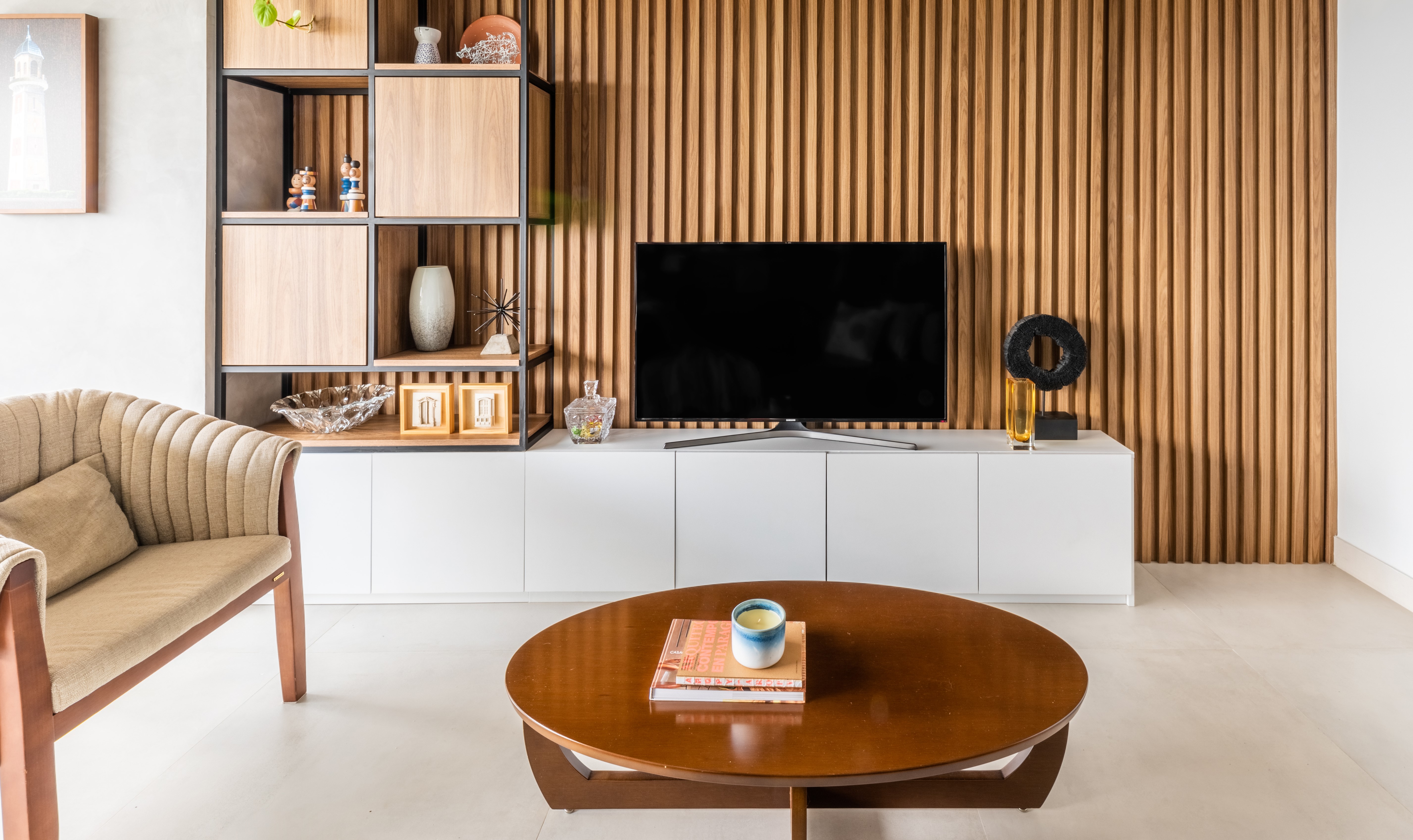 Sala de estar moderna com parede ripada de madeira clara, estante com nichos e arm&aacute;rio branco. Uma TV preta est&aacute; sobre o arm&aacute;rio, e uma mesa de centro redonda de madeira escura tem livros e uma vela. Um sof&aacute; bege com almofada est&aacute; &agrave; esquerda