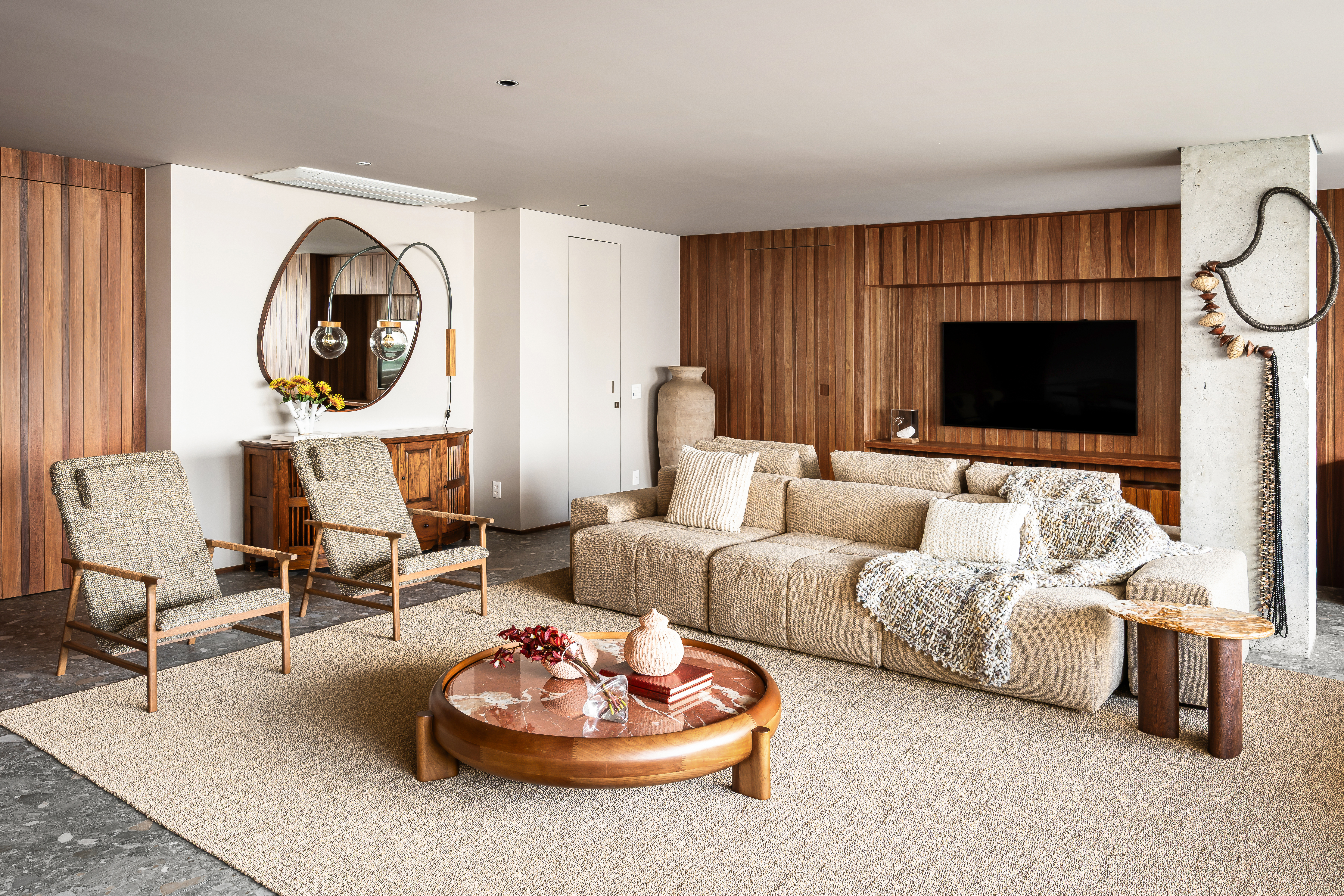 Sala de estar com sof&aacute; modular bege, duas poltronas de madeira e tecido claro, e uma mesa de centro redonda de madeira com tampo de m&aacute;rmore. A parede principal tem pain&eacute;is de madeira e uma TV, enquanto outra parede exibe um espelho org&acirc;nico e um aparador r&uacute;stico com flores amarelas. Um tapete bege cobre o ch&atilde;o, e uma escultura abstrata decora uma coluna de concreto