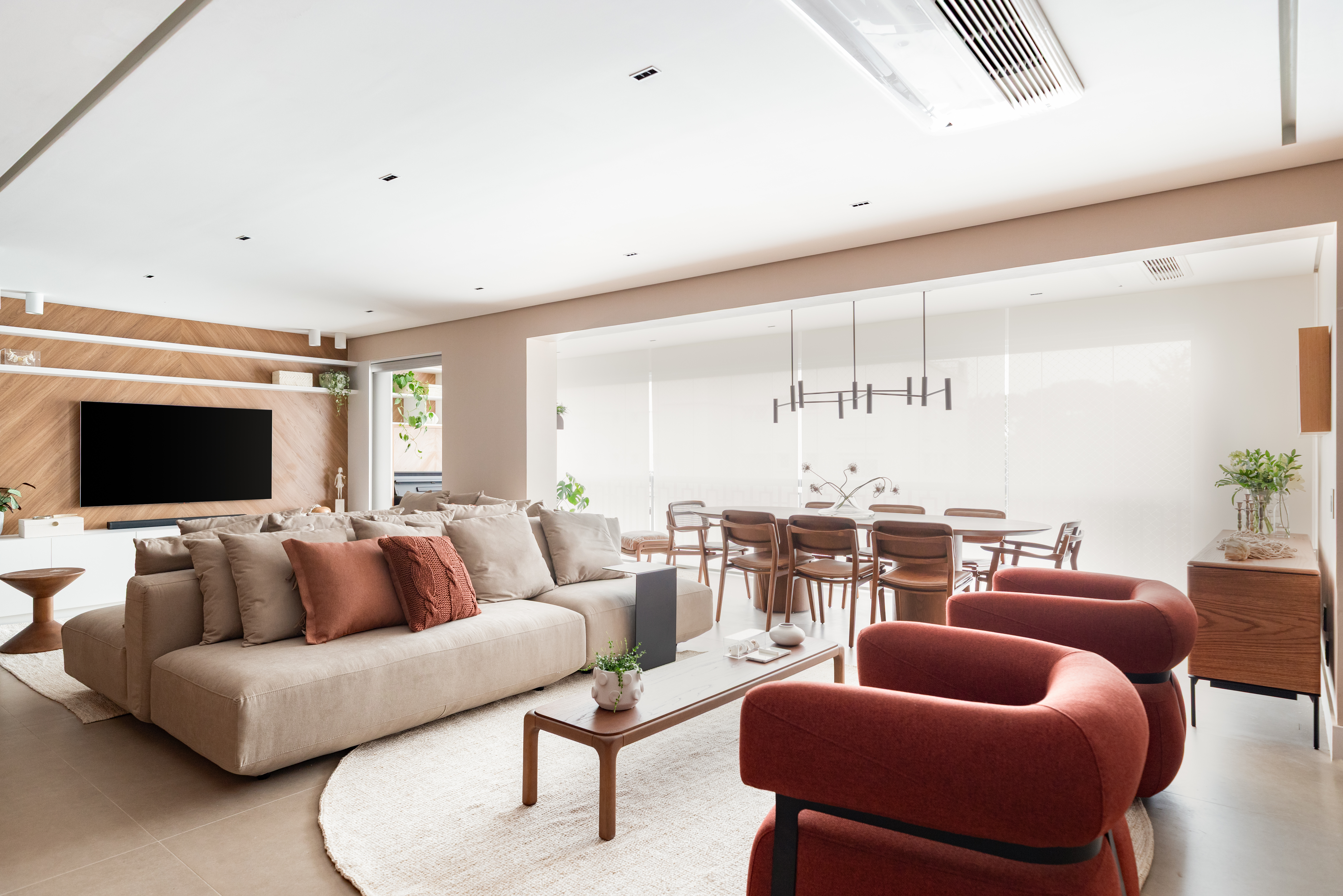 Sala de estar e jantar integradas com sof&aacute; bege, almofadas terracota, poltronas vermelhas e mesa de centro de madeira. Ao fundo, painel de TV amadeirado e mesa de jantar com cadeiras de madeira. O ambiente &eacute; iluminado e moderno